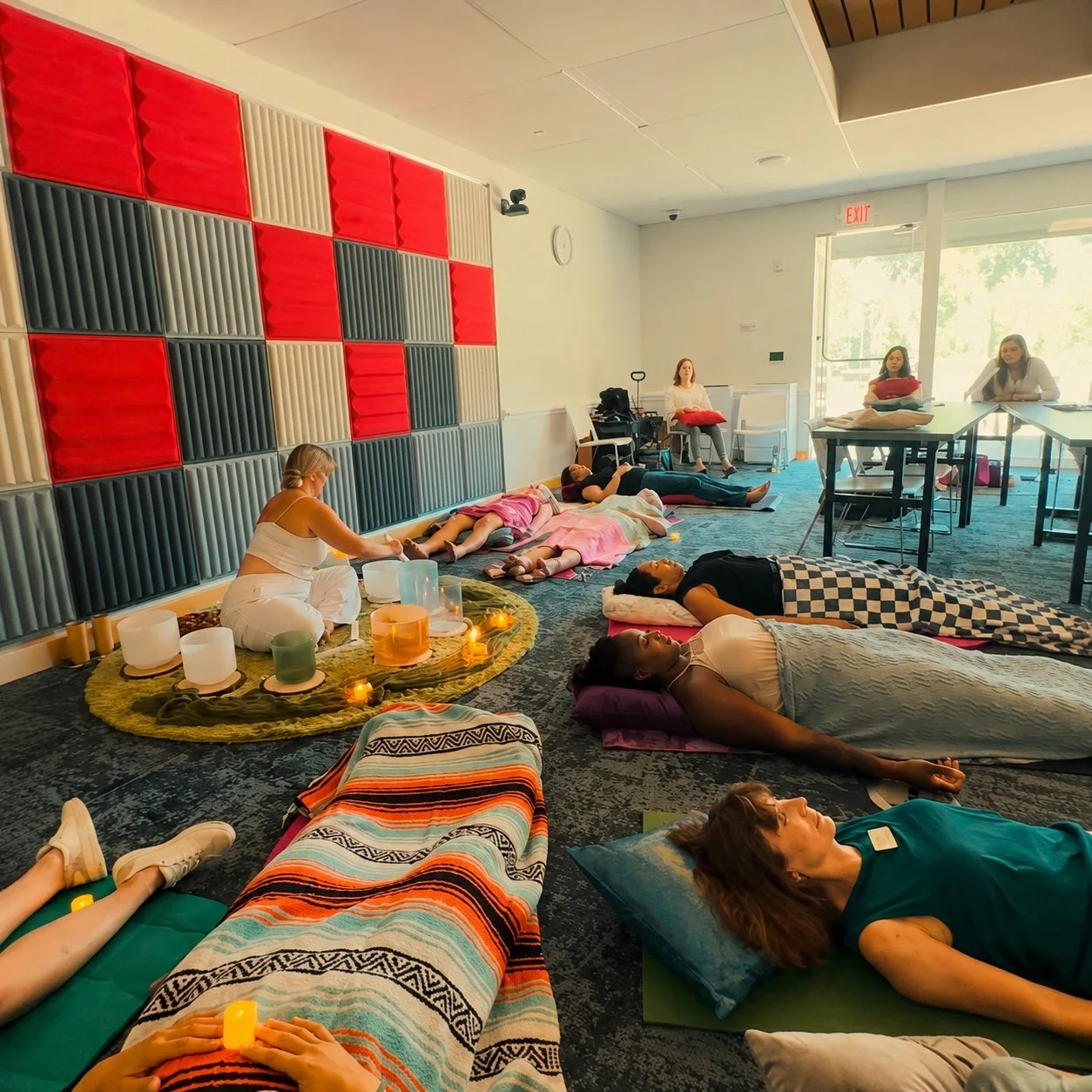 People participating in a sound bath meditation session, lying on mats and blankets in a room with acoustic panels on the wall, some holding candles, with sunlight coming through large windows.
