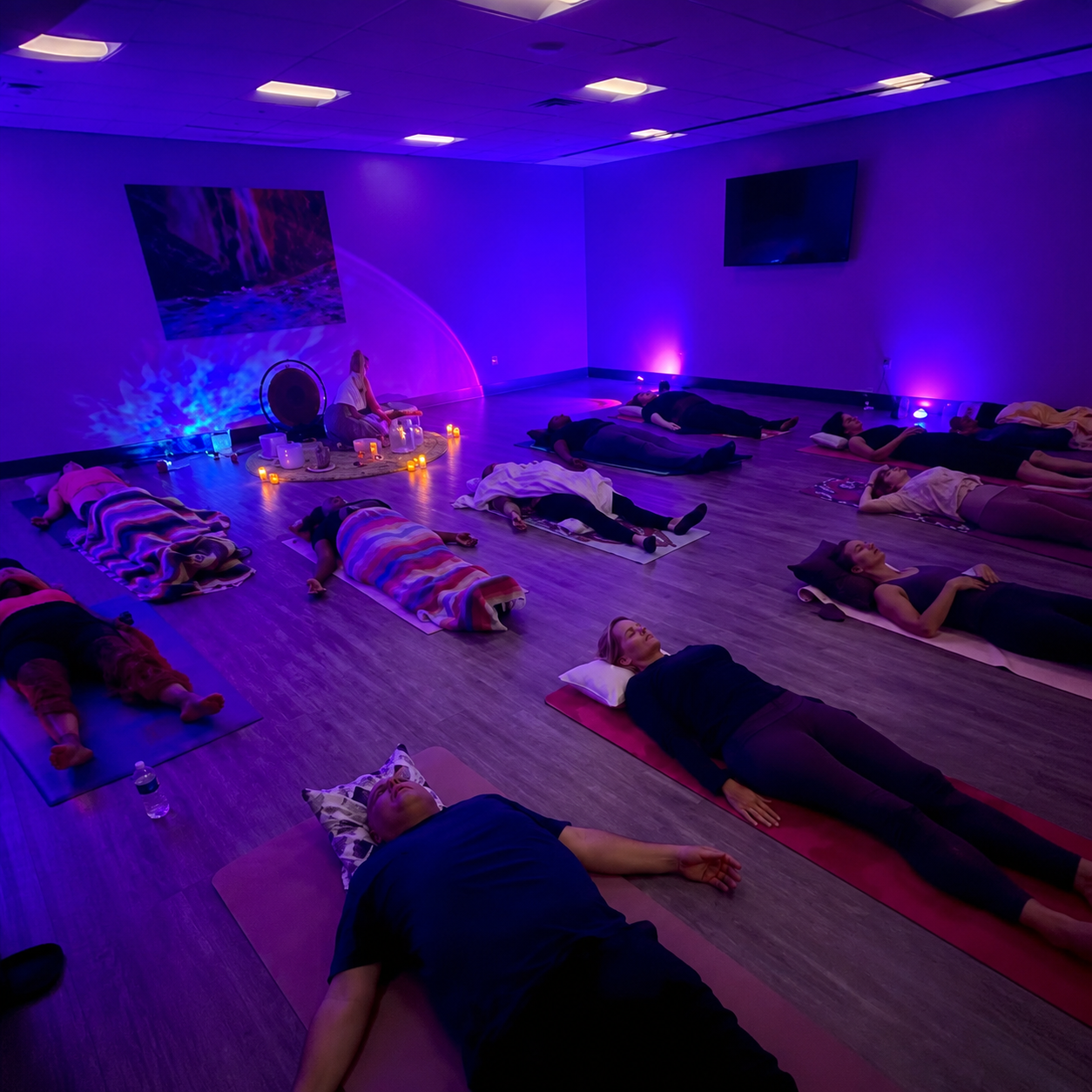 People participating in a yoga or meditation class in a dimly lit room with purple and blue lighting, lying on mats with some covered with blankets, candles, and candles around a instructor at the front, with a large screen and artwork on the walls.