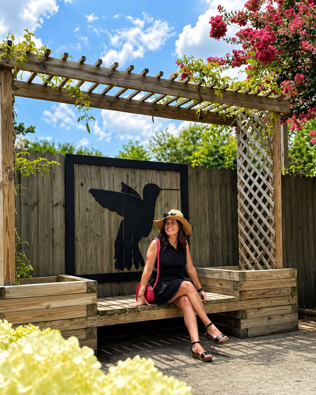 A woman sitting on a wooden bench under a pergola with green vines, wearing a wide-brimmed hat and a black dress, smiling at a garden area with blooming flowers and a backyard fence with a hummingbird silhouette in the background.