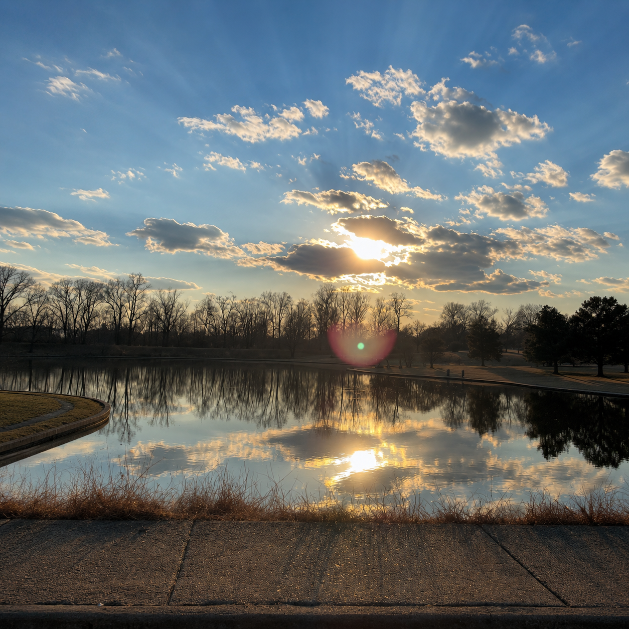 Sunset over a calm lake with bare trees reflected in the water, partly cloudy sky, and a sidewalk in the foreground.