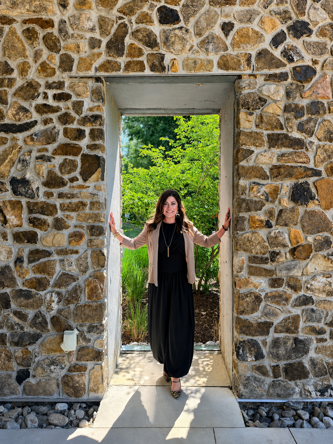 A woman standing in a stone doorway, smiling, with greenery behind her on a sunny day.