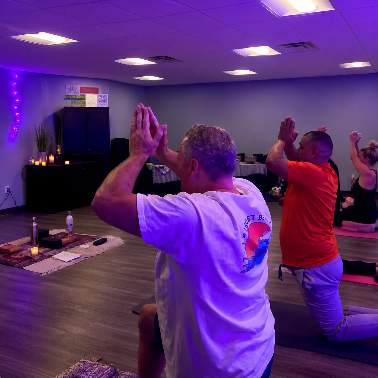 People practicing yoga in a dimly lit room with purple lighting, candles, and a peaceful ambiance.