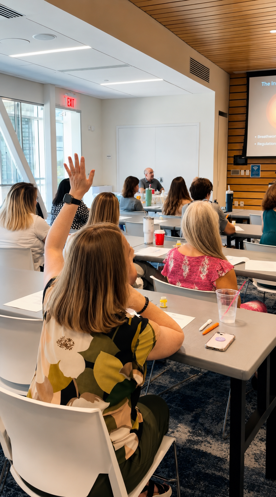 A classroom or conference room filled with people attending a presentation; one woman is raising her hand. There are papers, pens, and drinks on the tables, and a large screen displaying a presentation at the front.