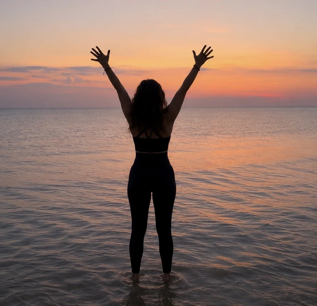 A woman standing in shallow water at the beach during sunset, with arms raised.