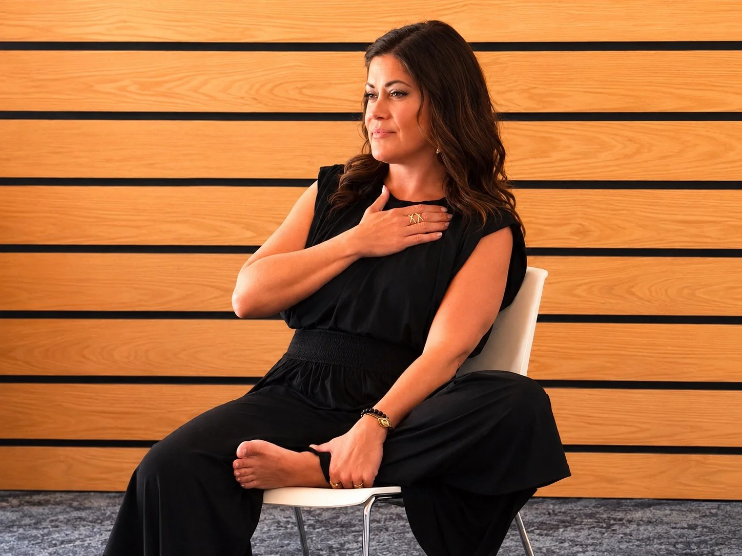 A woman in a black outfit sitting on a white chair against a wooden wall, with her right hand on her chest and her left hand holding her foot, appearing to be in a relaxed or contemplative pose.