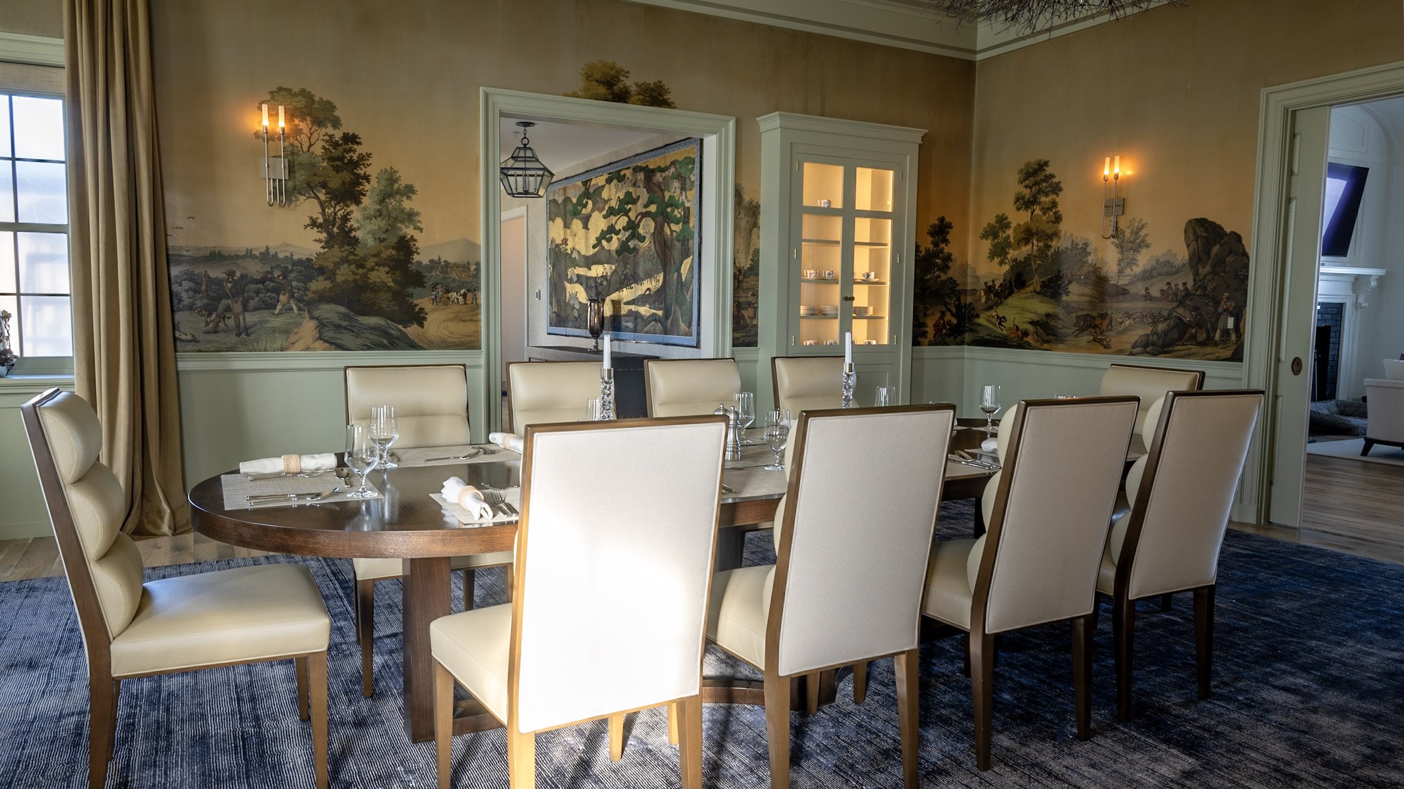 A formal dining room with a large wooden round table set with napkins, glasses, and candlesticks. Cream-colored chairs surround the table. The walls feature a scenic mural of trees, hills, and people. There is a built-in cabinet with glass doors disp