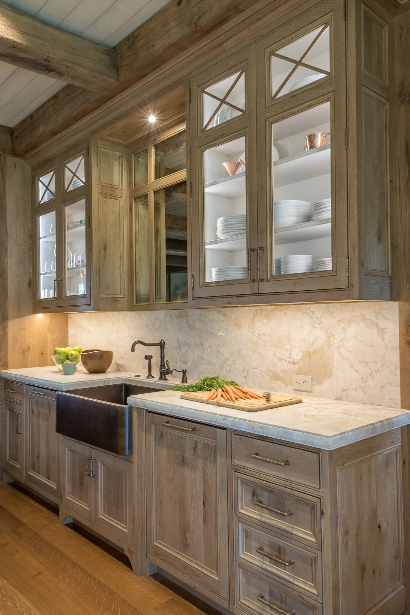 Rustic wooden kitchen cabinets with glass doors displaying dinnerware and cookware, marble countertop with a cutting board, carrots, and kitchen sink with faucet.