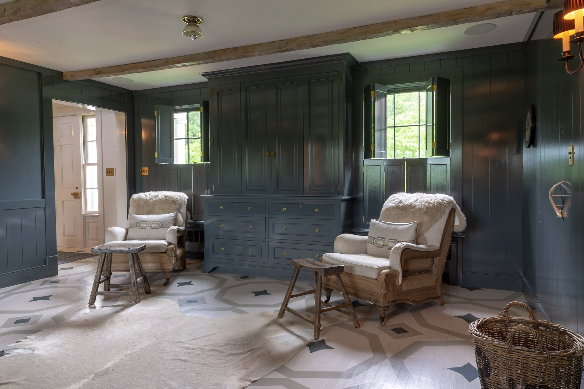 Living room with dark green paneled walls, two white armchairs with fur throws, small wooden side tables, a large blue cabinet, open window with green foliage visible, ceiling light fixture, patterned tile floor, and a wicker basket in the foreground