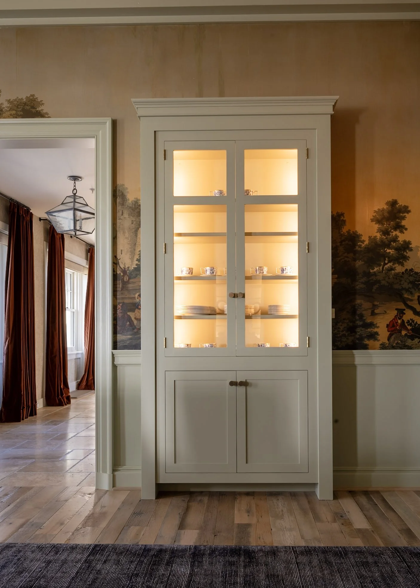 A white display cabinet with glass doors illuminated from within, showcasing dishes and small bowls, in a room with wooden floor and mural wallpaper with trees and people, and a separate room visible through an open doorway with curtains and a geomet