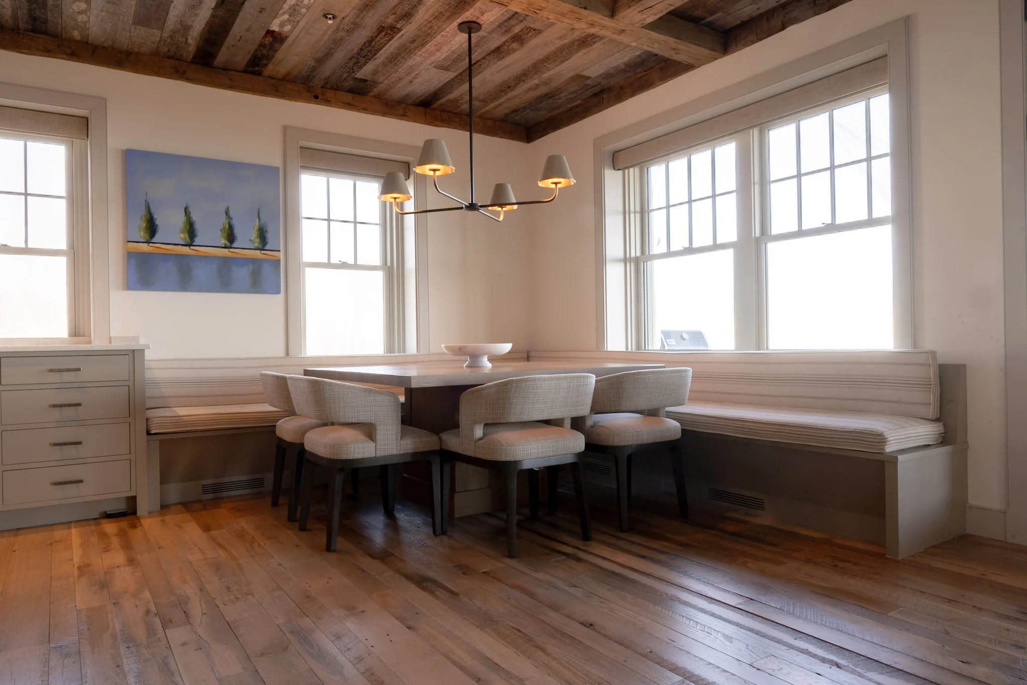 A dining area with a wooden ceiling and large windows, featuring a dining table with beige upholstered chairs, a chandelier with five lampshades, a painting on the wall depicting trees, and a built-in window seat with striped cushion.