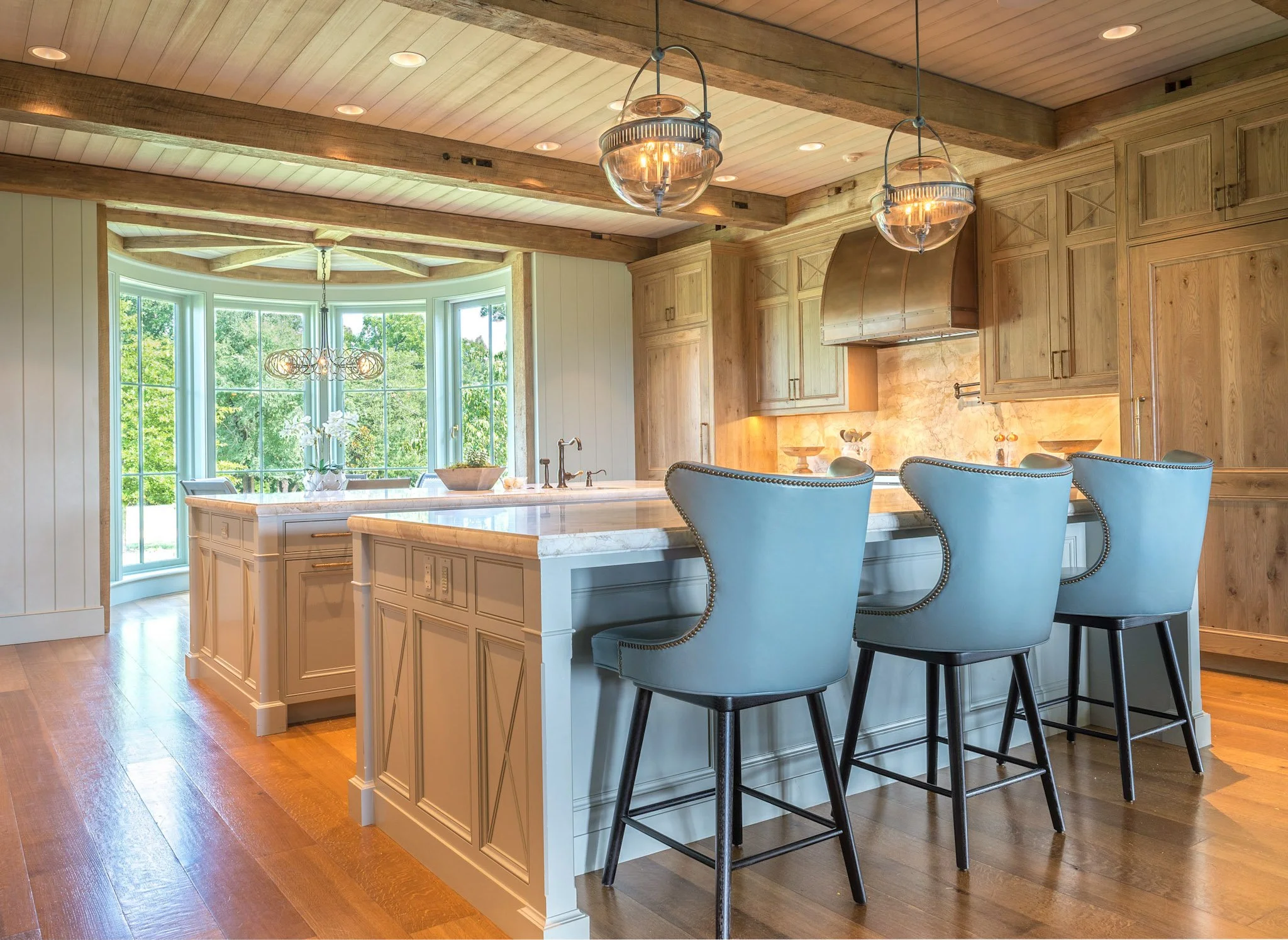 Inside a spacious kitchen with a large white island, four blue chairs with dark legs around the island, wooden cabinetry, a range hood, large windows with scenic green outdoor view, and pendant lights hanging from a wooden ceiling.