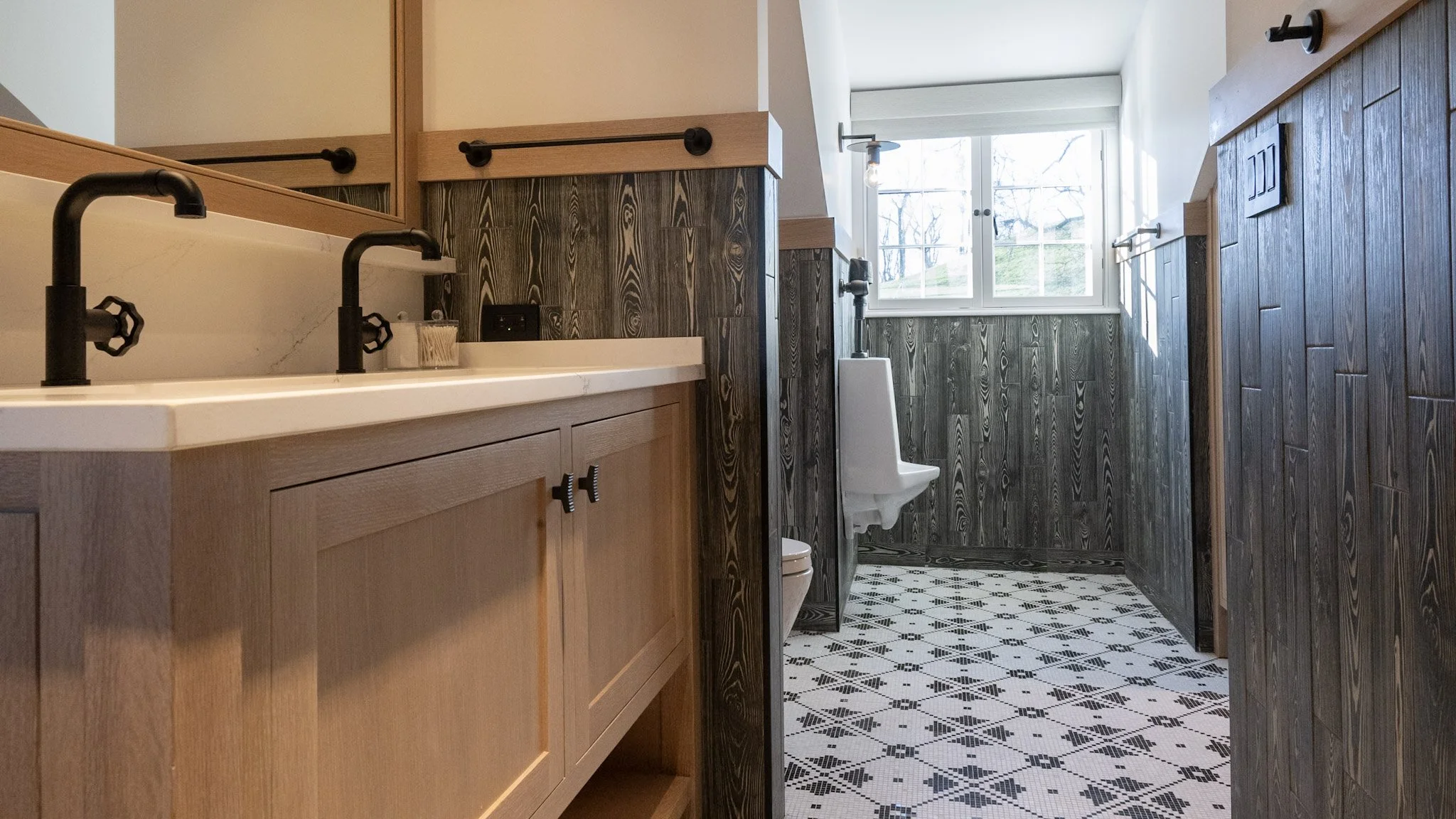 A bathroom with a wooden vanity, black faucets, a tiled wall, a urinal, and a window with a view of trees outside.