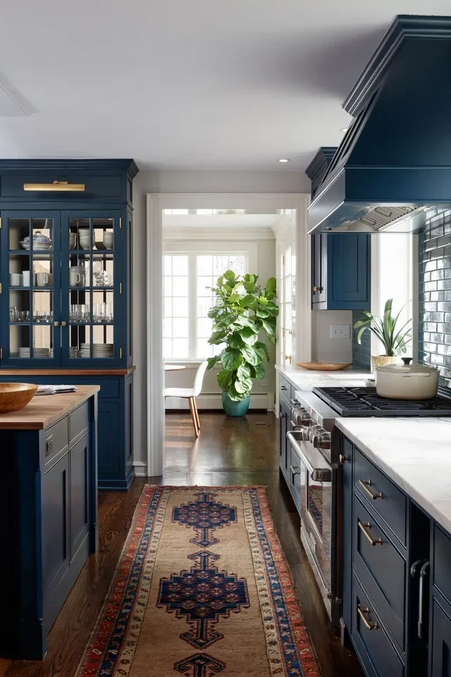 Kitchen with dark blue cabinets, white marble countertops, and a colorful runner rug on dark hardwood floors. There is a large leafy green plant near a window at the end of the room.
