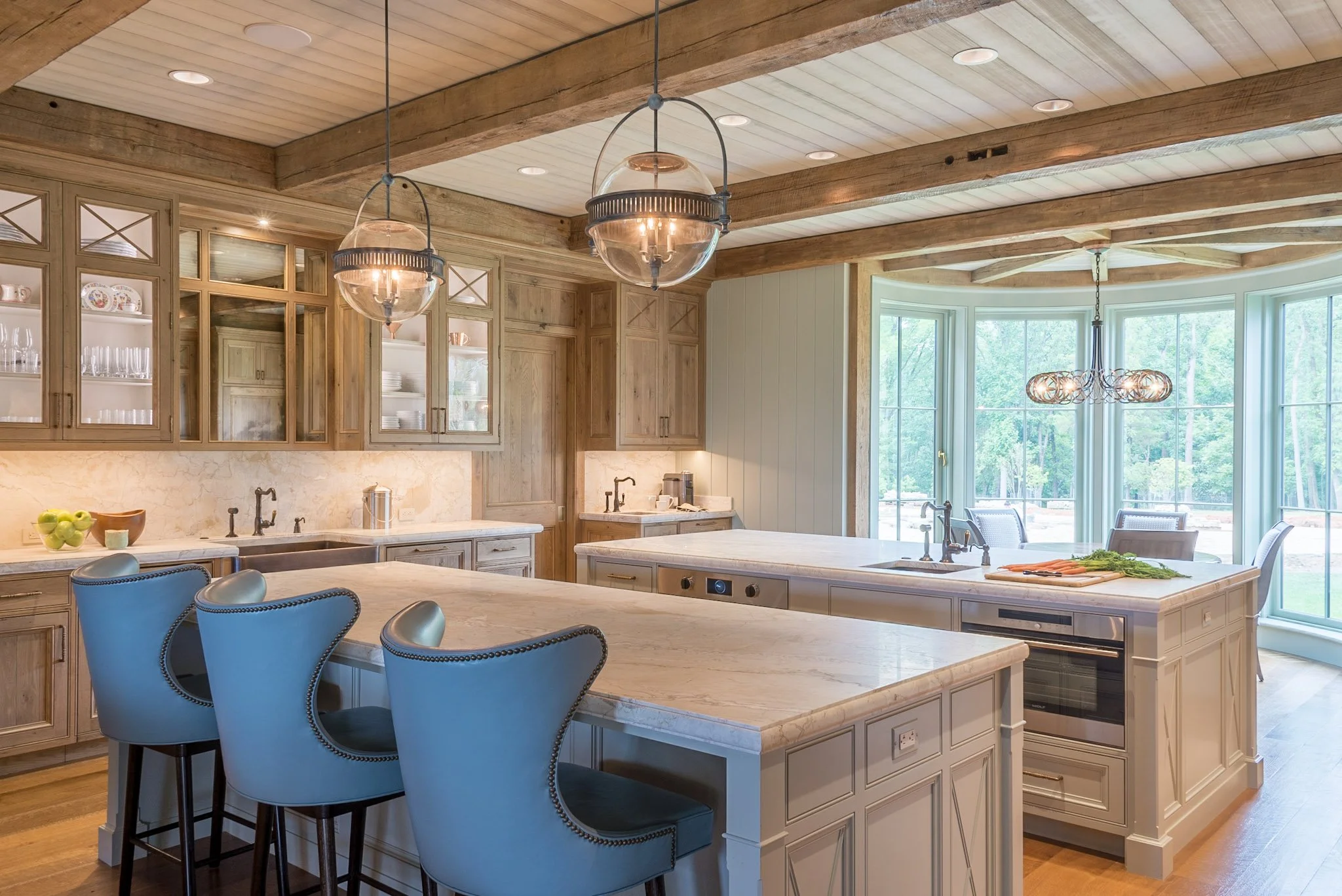 Kitchen with light wood cabinetry, marble countertops, and a large center island. There are two spherical pendant lights hanging from the ceiling, which has wooden beams. A breakfast bar with upholstered chairs is visible in the foreground. Large win