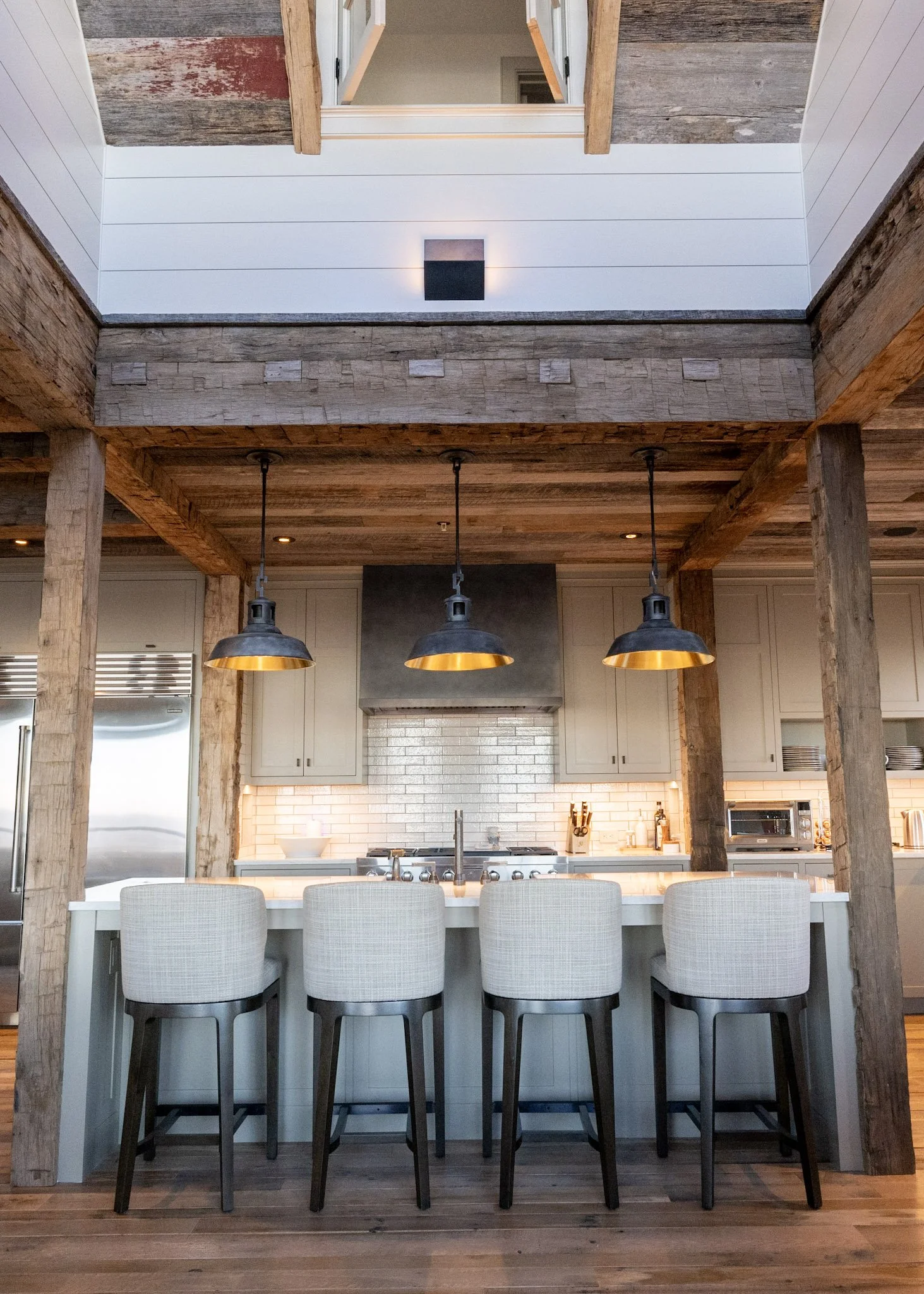 A remodeled modern kitchen with a white upholstered bar stools, wooden beams, and hanging pendant lights.