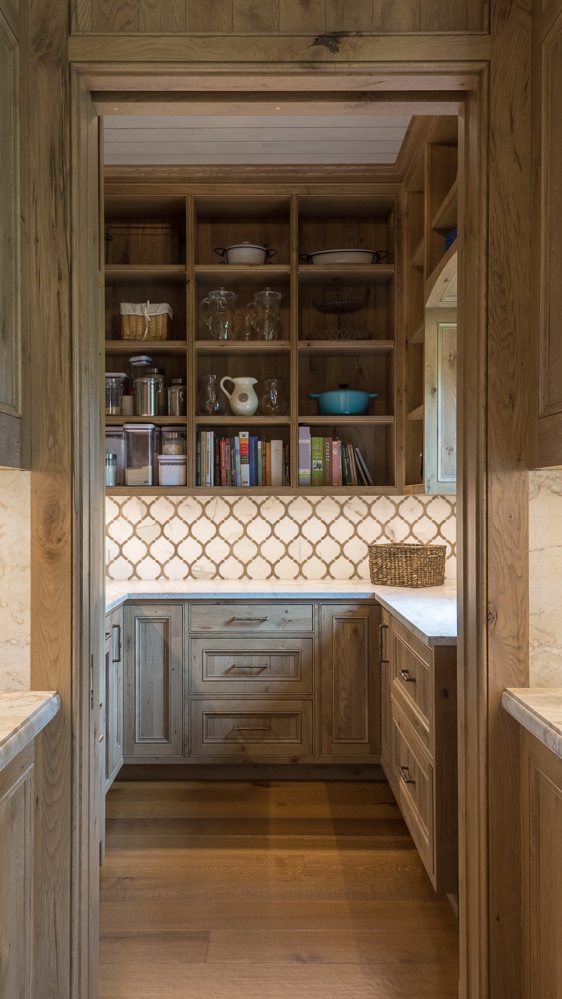 A kitchen cabinet with open shelves displaying glassware, cookbooks, and kitchenware, with a decorative backsplash and a basket on the counter.