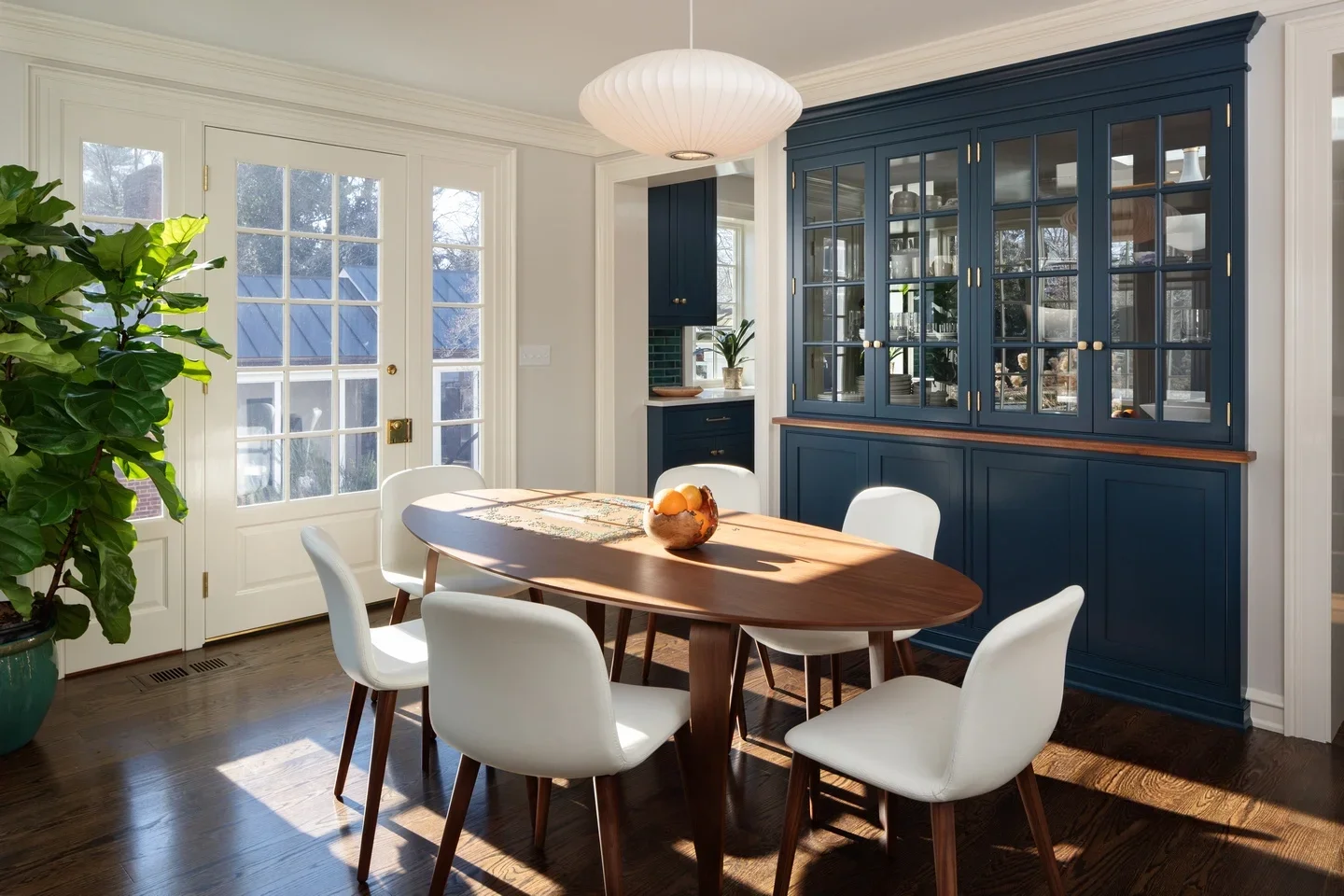 A dining room with a wooden table surrounded by six white chairs, a blue china cabinet, glass-paneled double doors, a large green potted plant, and sunlight streaming through the windows.