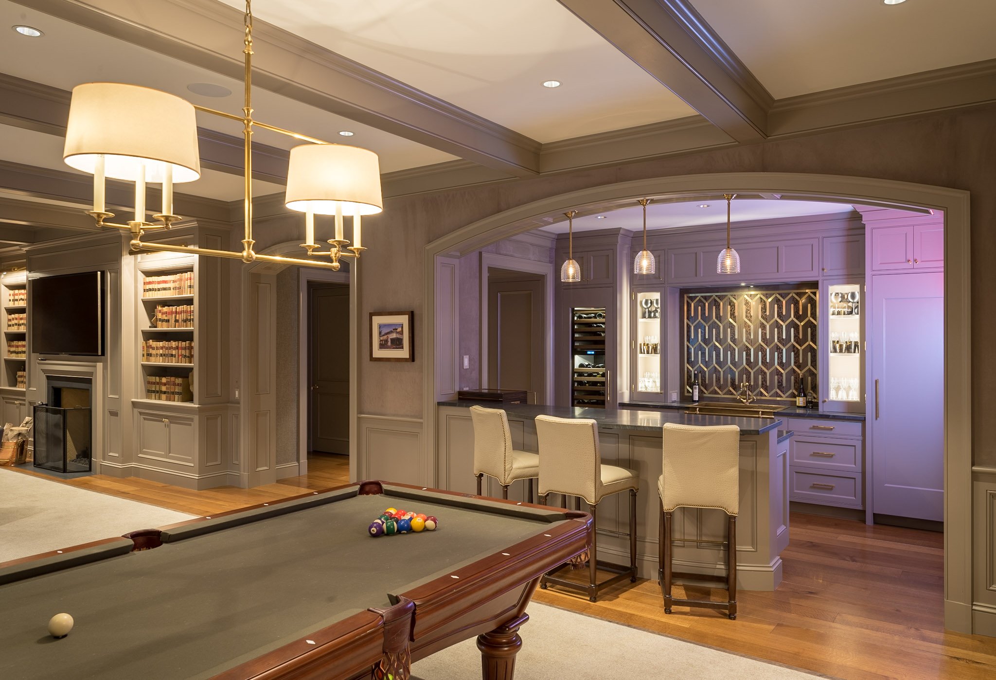 A luxurious living area featuring a pool table with balls set up, a bar area with purple lighting, bookshelves with books, and elegant ceiling lighting fixtures.