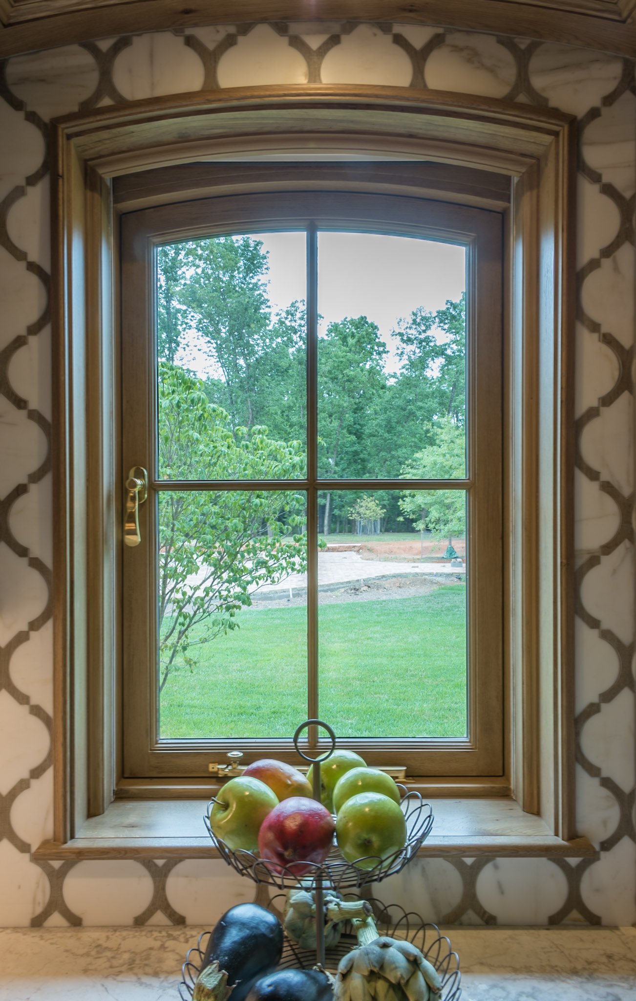 A kitchen window with a view of a green backyard with trees. On the windowsill, there is a two-tiered wire fruit basket filled with apples, garlic, and eggplants.