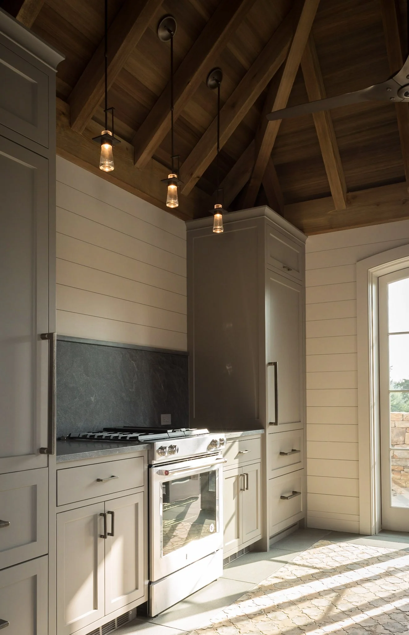 A modern kitchen with white cabinetry, a gray countertop, a stainless steel stove, and a wooden vaulted ceiling with pendant lights and a ceiling fan.