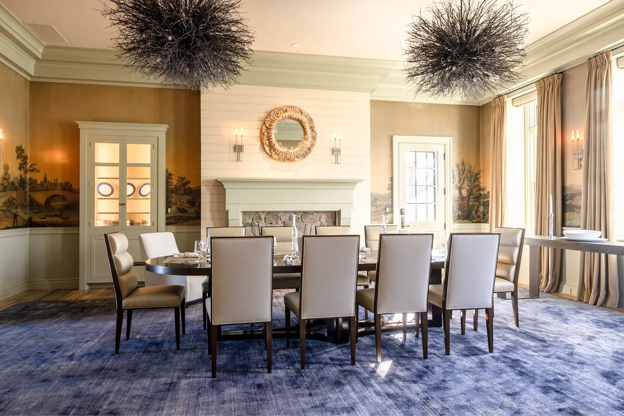 Elegant dining room with a large table surrounded by cream-colored chairs, a white brick fireplace with a decorative mirror above, landscape murals on the walls, large windows with curtains, and two black chandeliers hanging from the ceiling.