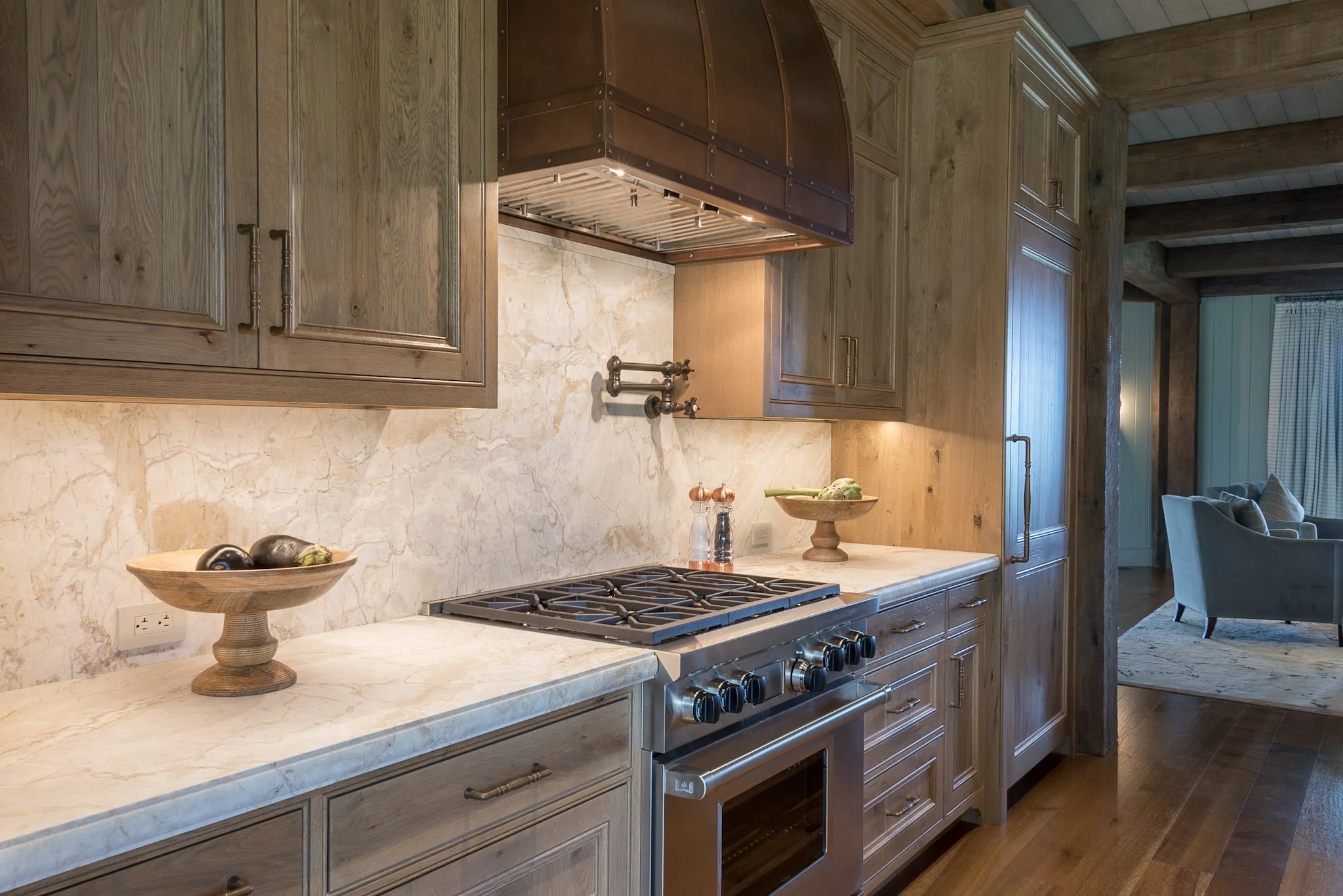 A rustic kitchen with wooden cabinets and a marble countertop. There is a stove with oven, decorative bowls with vegetables, and a small window with blue curtains in the background.