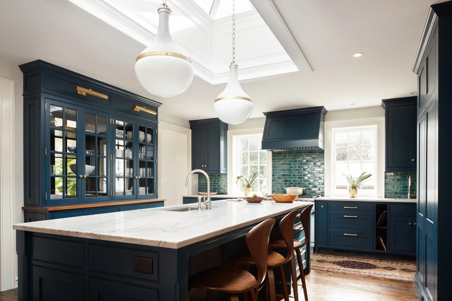 A modern kitchen with navy blue cabinets, a white marble island, and pendant lighting. Features a green tiled backsplash, wooden chairs, plants near windows, and a patterned rug on wooden floors.