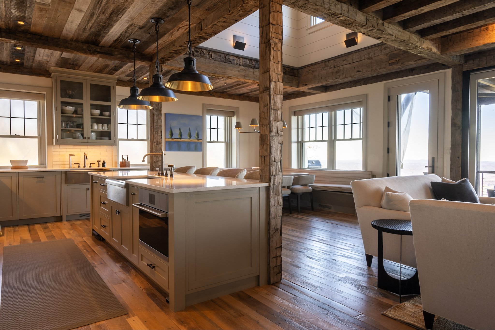 Open-concept kitchen and living room with rustic wood beams, large windows, and modern pendant lighting, featuring a kitchen island with a sink and white cabinetry, and a cozy seating area with a beige sofa.