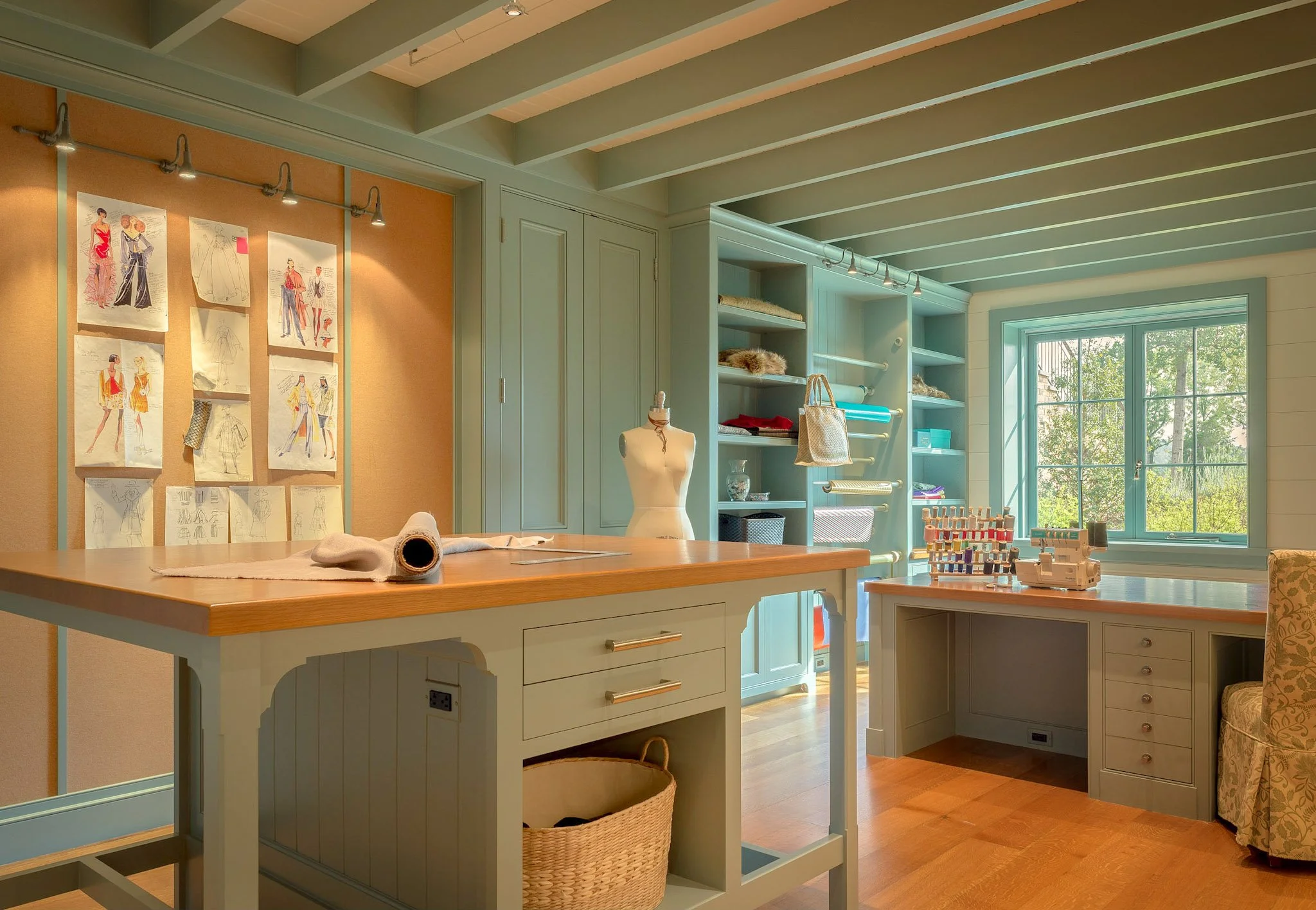 A bright sewing or craft room with a large window letting in natural light. The room features mint-colored cabinetry, open shelves with fabric and supplies, and a cutting table with sewing tools. Sewing pattern sketches are pinned on the wall, and a 