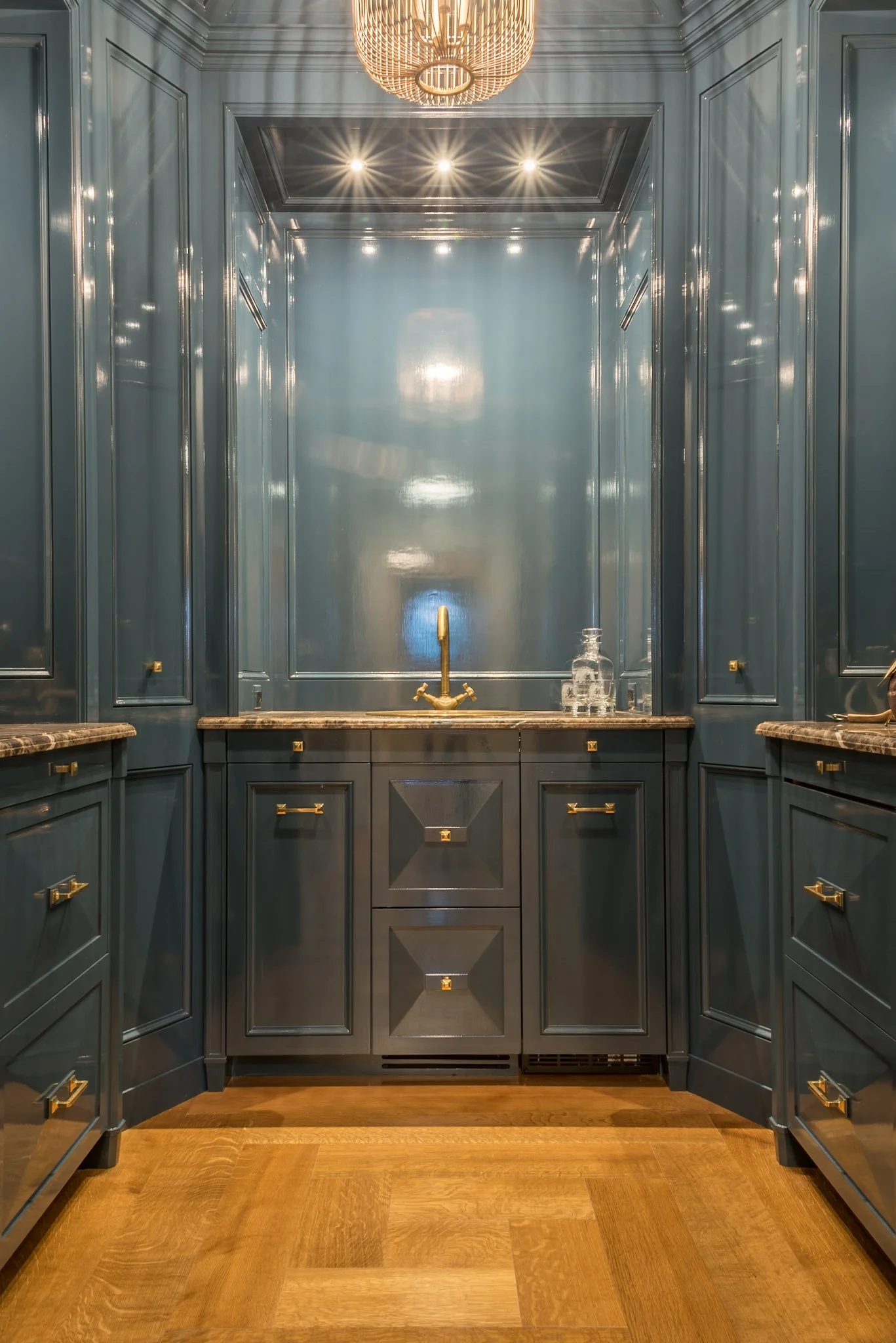 A luxurious kitchen with dark blue cabinets featuring gold hardware. The back wall has a gold faucet and a marble countertop with a glass jar. A modern chandelier hangs from the ceiling, and bright recessed lights illuminate the space. The floor is m