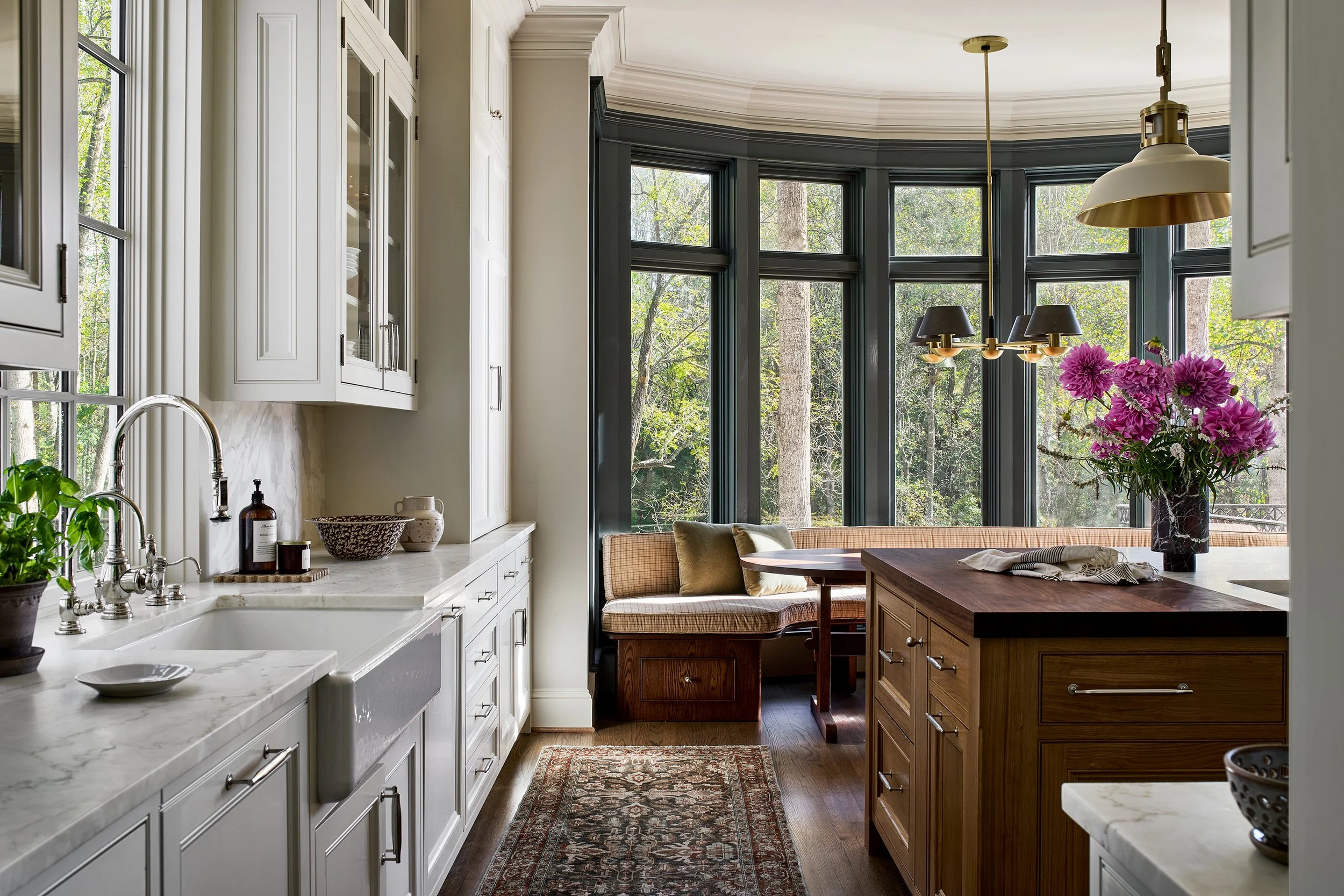 A cozy kitchen with white cabinets, a marble countertop, and a wooden island. Large windows with black trim offer a view of trees outside. A round wooden table with a pink floral arrangement and a woven bench are visible, along with hanging light fix