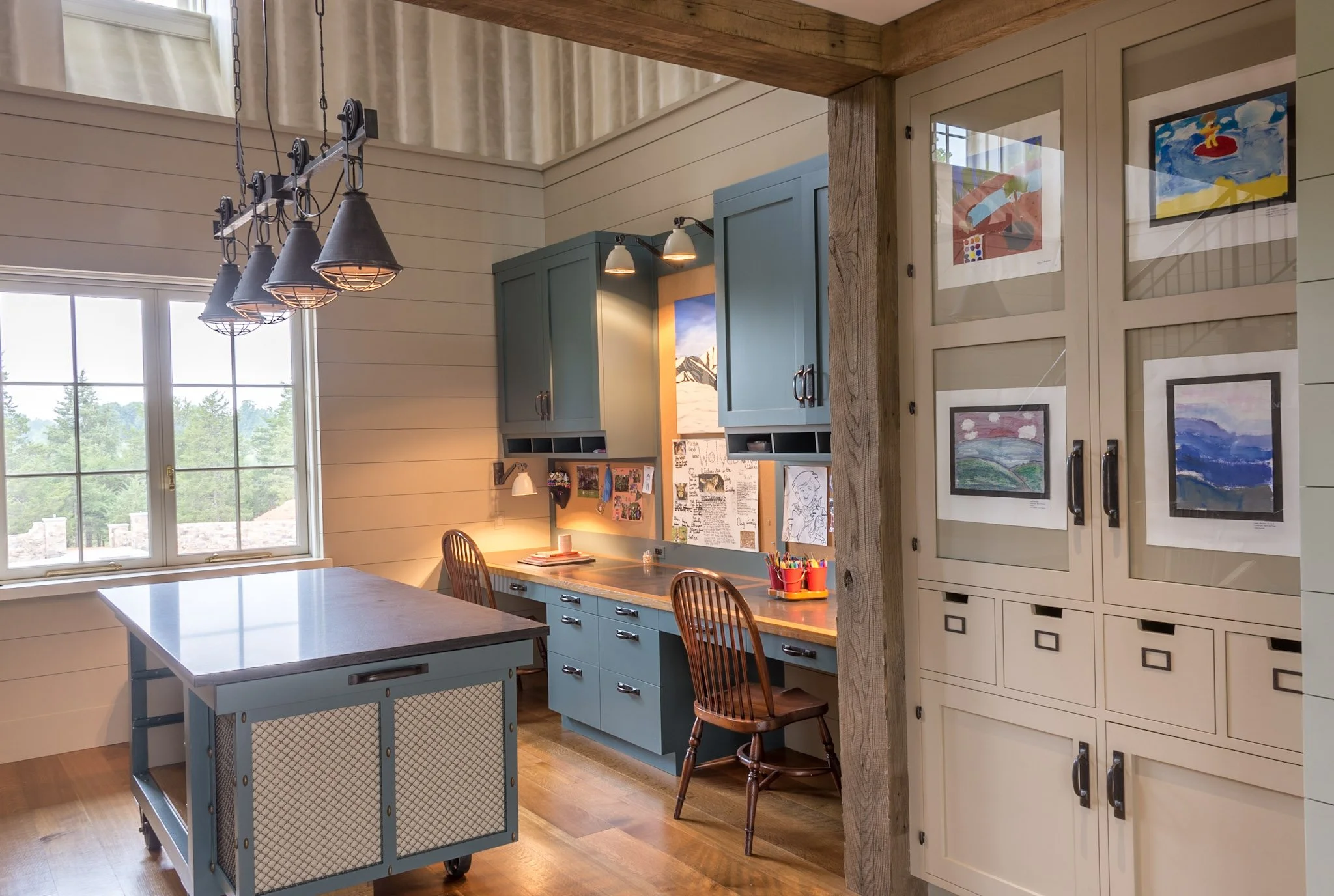A cozy kitchen with large window, green cabinetry, and a dining table with chairs. There are colorful children's drawings and artwork on the cabinets and wall. A black metal chandelier hangs above the counter, which is illuminated by natural and inst