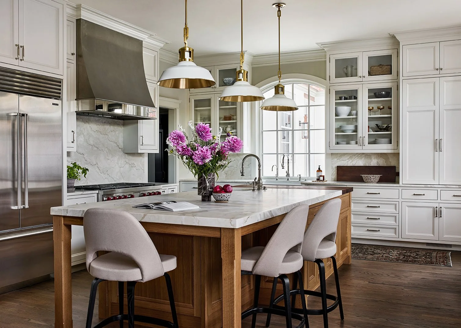 A modern kitchen with white cabinets, a marble island with a wooden base, and three pendant lights with gold accents. There are purple flowers in a vase, a bowl of apples, and a book on the island. A large window lets in natural light.
