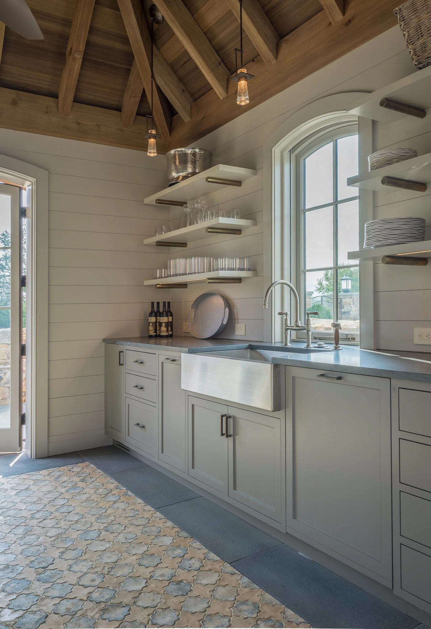 A country-style kitchen with white cabinetry, a farmhouse sink, open floating shelves with dishes and glasses, a window with a view outside, and textured tile flooring. Exposed wooden ceiling beams and pendant lights add rustic charm.