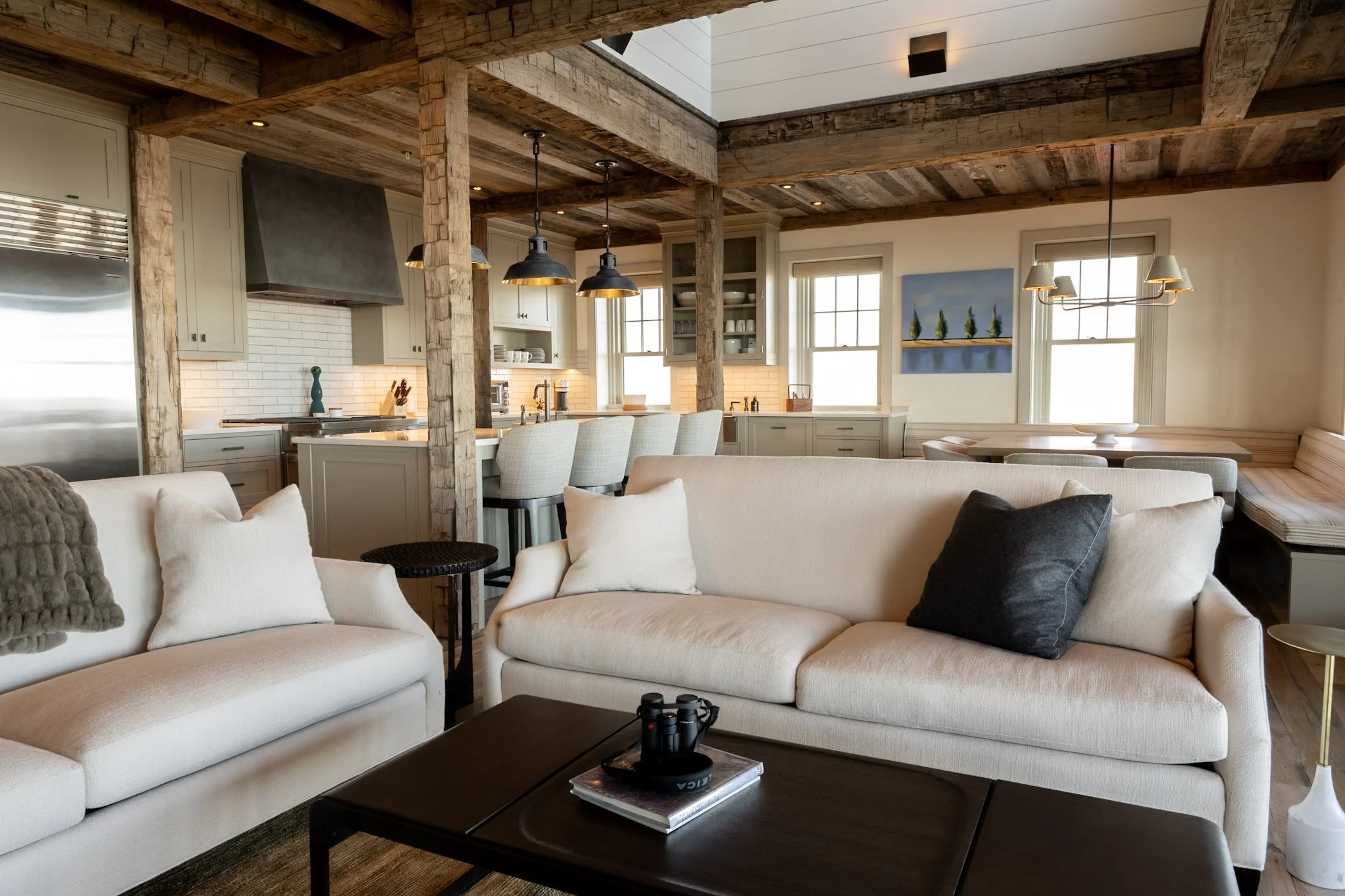 Living room with white couches, pillows, and a coffee table with binoculars and books, open kitchen with pendant lights, white cabinetry, and wooden beams on the ceiling, large windows with blinds, and a painting on the wall.