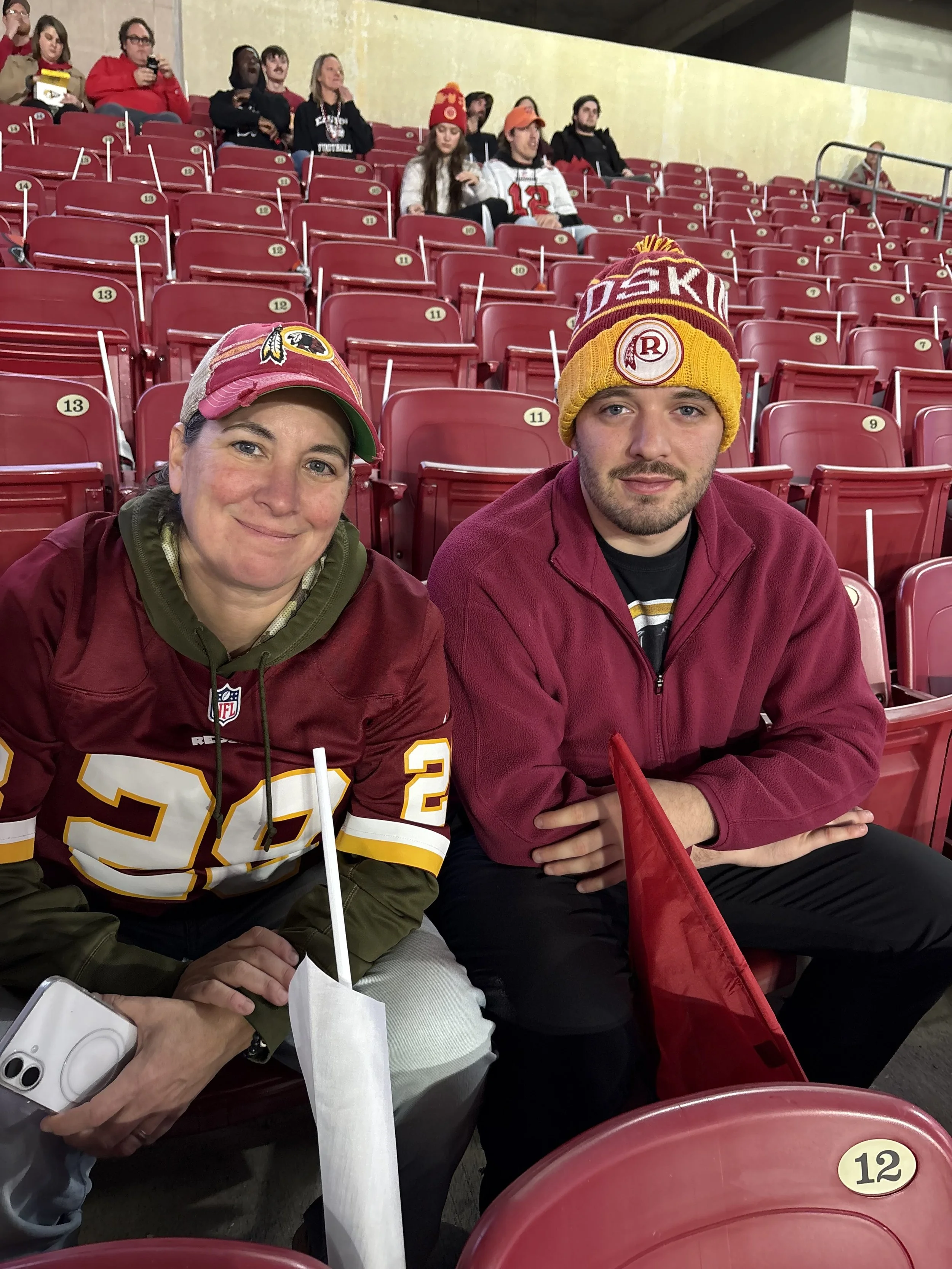 Two fans at a sports stadium sitting in red seats, wearing football merchandise, including hats and jerseys of the Washington Football Team, with other spectators in the background also watching the game.
