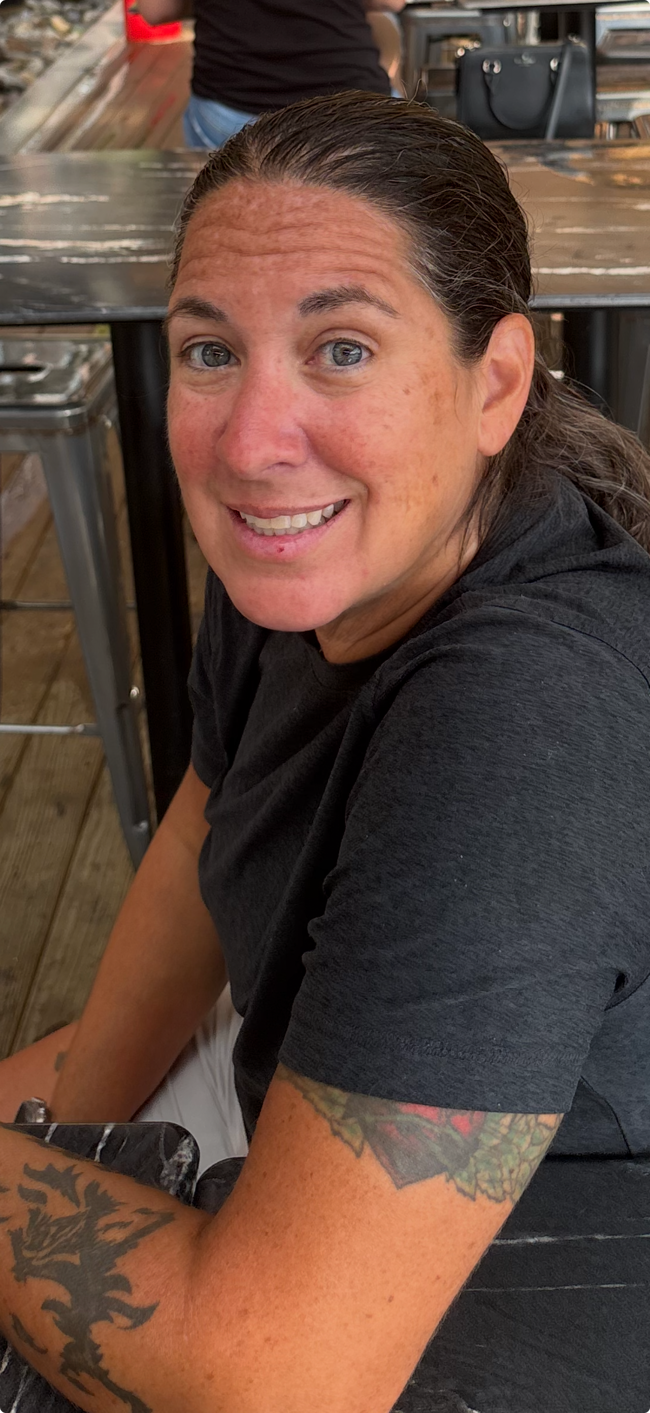 A woman with tattoos on her arms, smiling, sitting at a table in a casual setting.