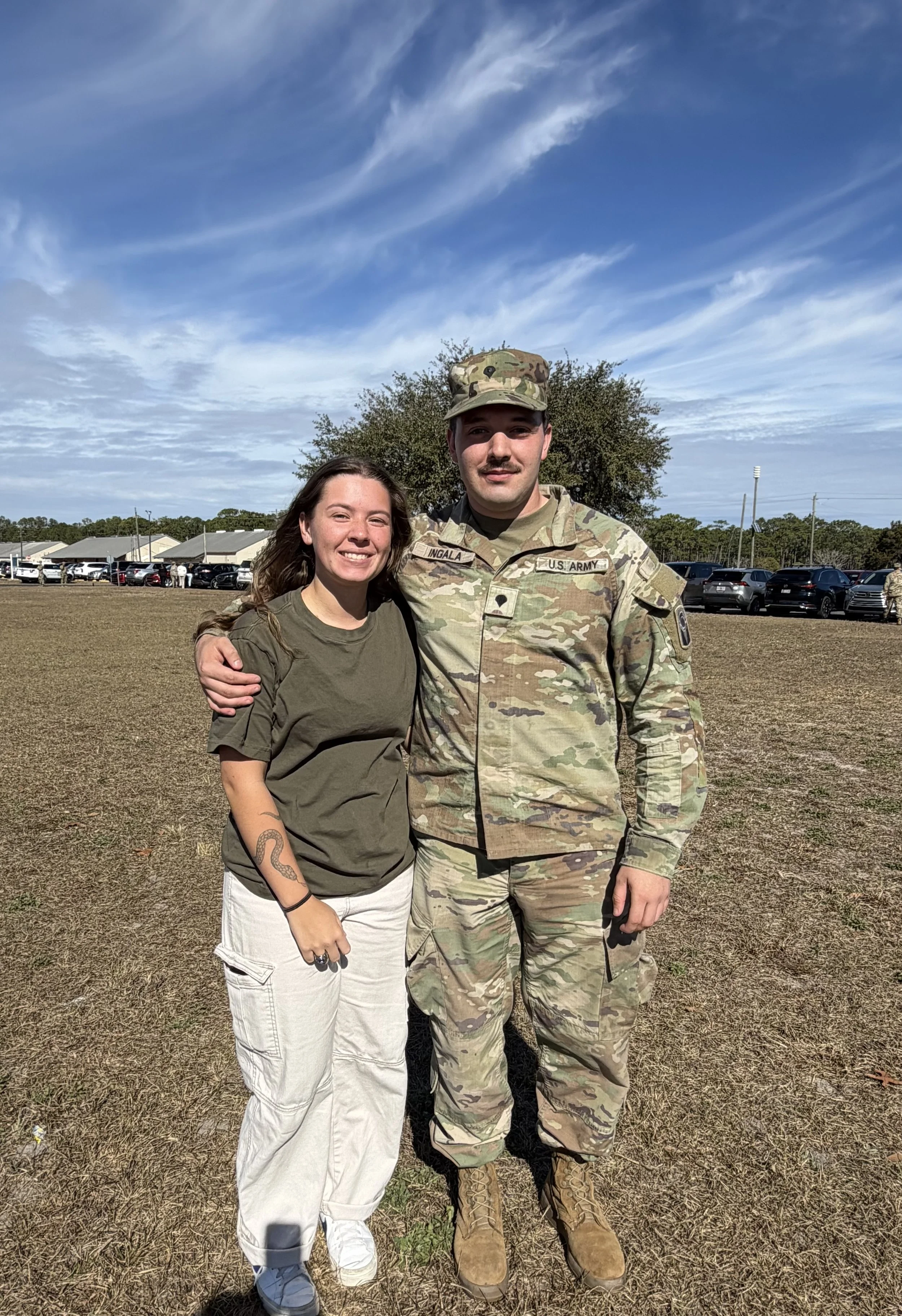 A woman and a soldier in uniform are standing outdoors, smiling, with their arms around each other. The woman has long brown hair, wears a green t-shirt and white pants, and displays a snake tattoo on her left forearm. The soldier is dressed in camouflage uniform and a cap, with a name tag reading