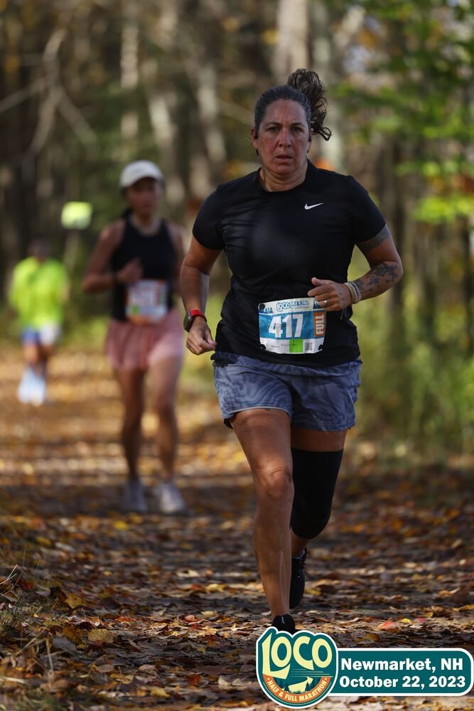 A woman running in a marathon through a forested trail during fall, wearing a black Nike shirt, shorts, a knee brace, and a race bib numbered 417, with two other runners visible in the background.