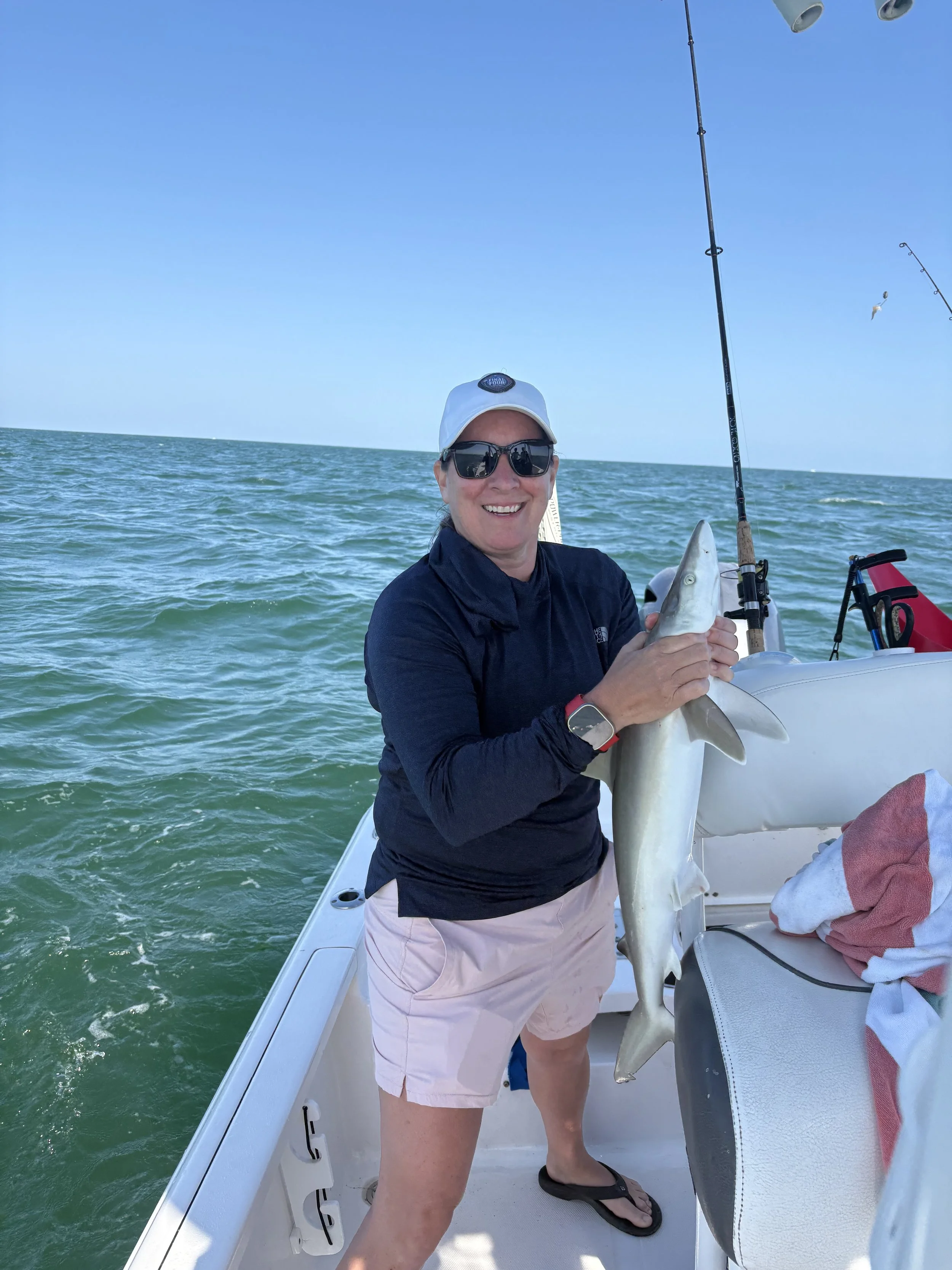 Woman on a boat holding a fish in her hand, wearing sunglasses, a white cap, black jacket, and light shorts, with fishing rods and ocean in the background.