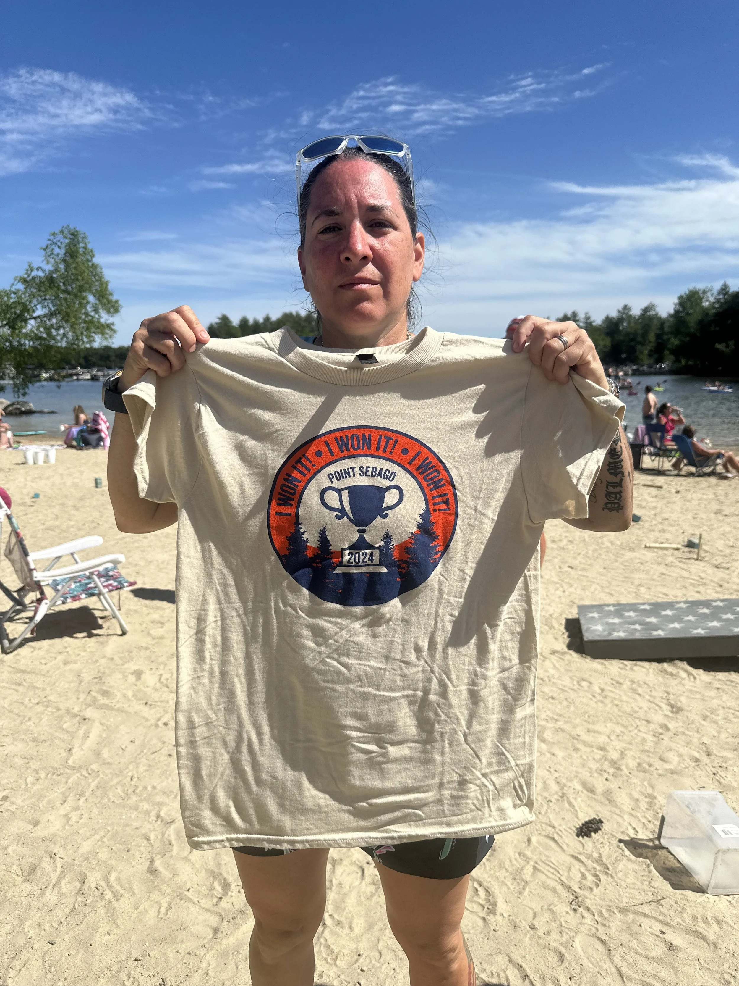 A woman on a sandy beach holding a beige T-shirt with the text 'Point Sebago' and 'I Won It! 2024' printed on it. She has sunglasses on her head, and there are other people, trees, and boats visible in the background under a partly cloudy blue sky.