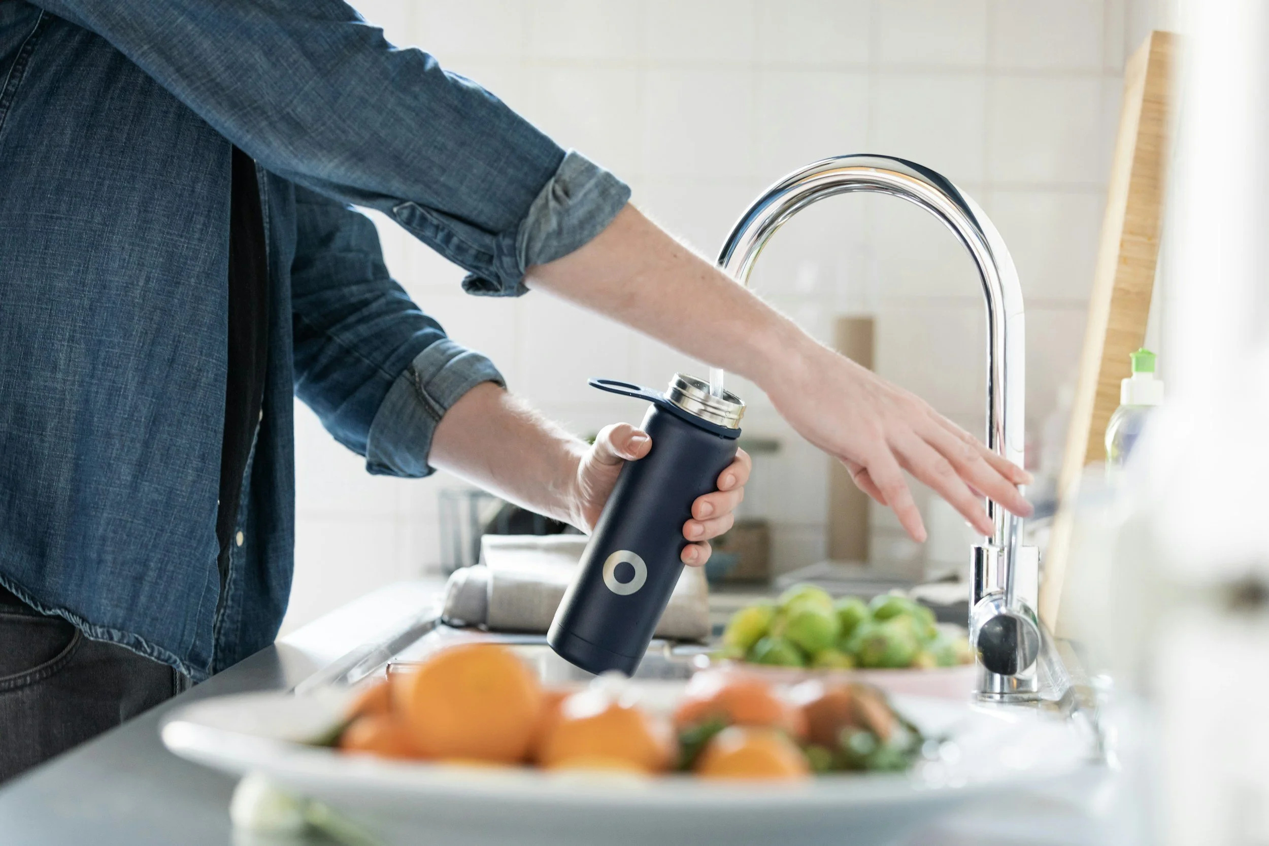 Person in denim shirt pouring water from a black reusable bottle into a kitchen sink.