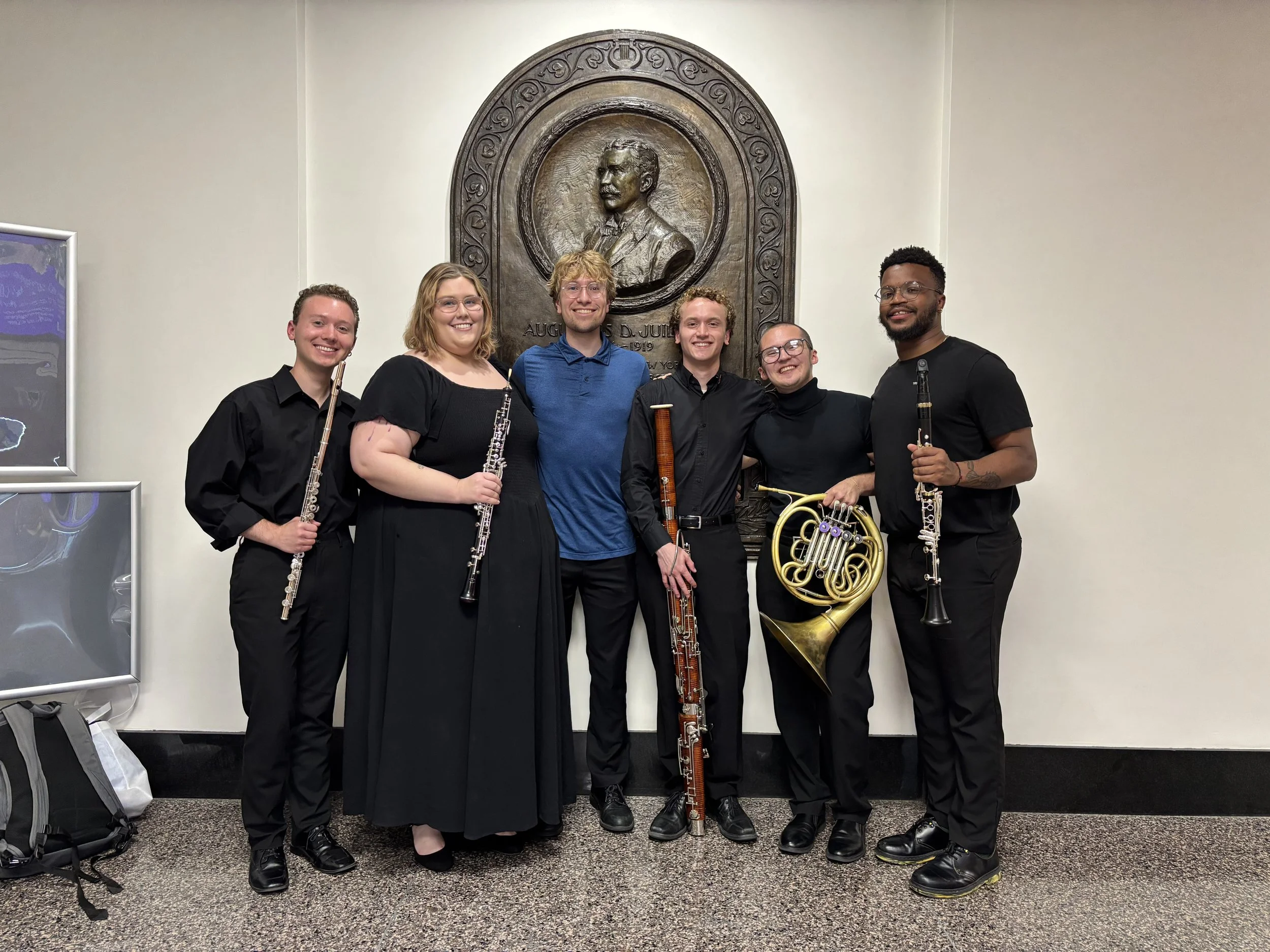 Six musicians holding musical instruments, standing in front of a bronze plaque of a man in a suit, in an indoor setting.