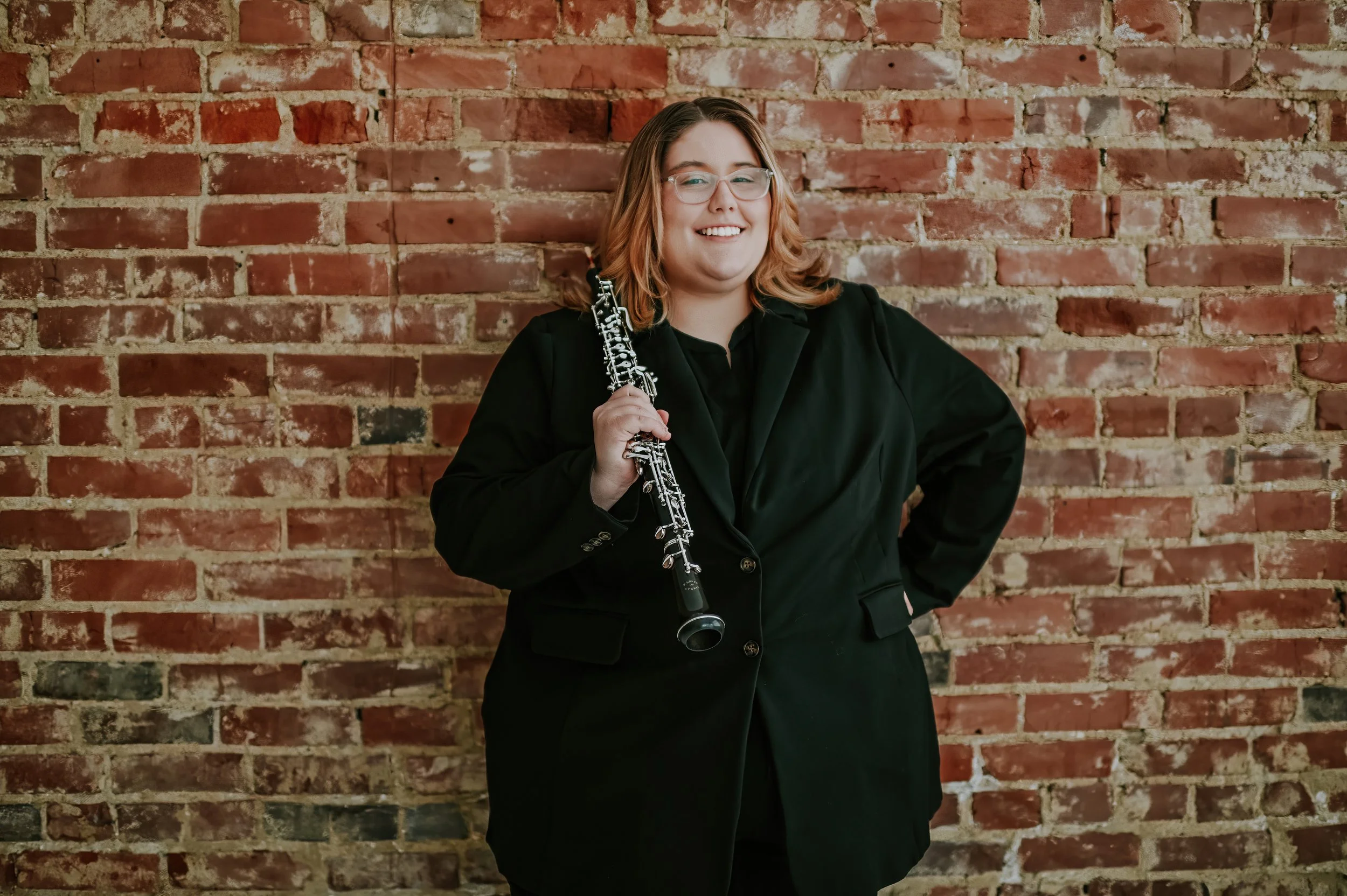 A woman with shoulder-length curly hair, wearing glasses and a black outfit, is smiling and holding a clarinet while standing in front of a brick wall.