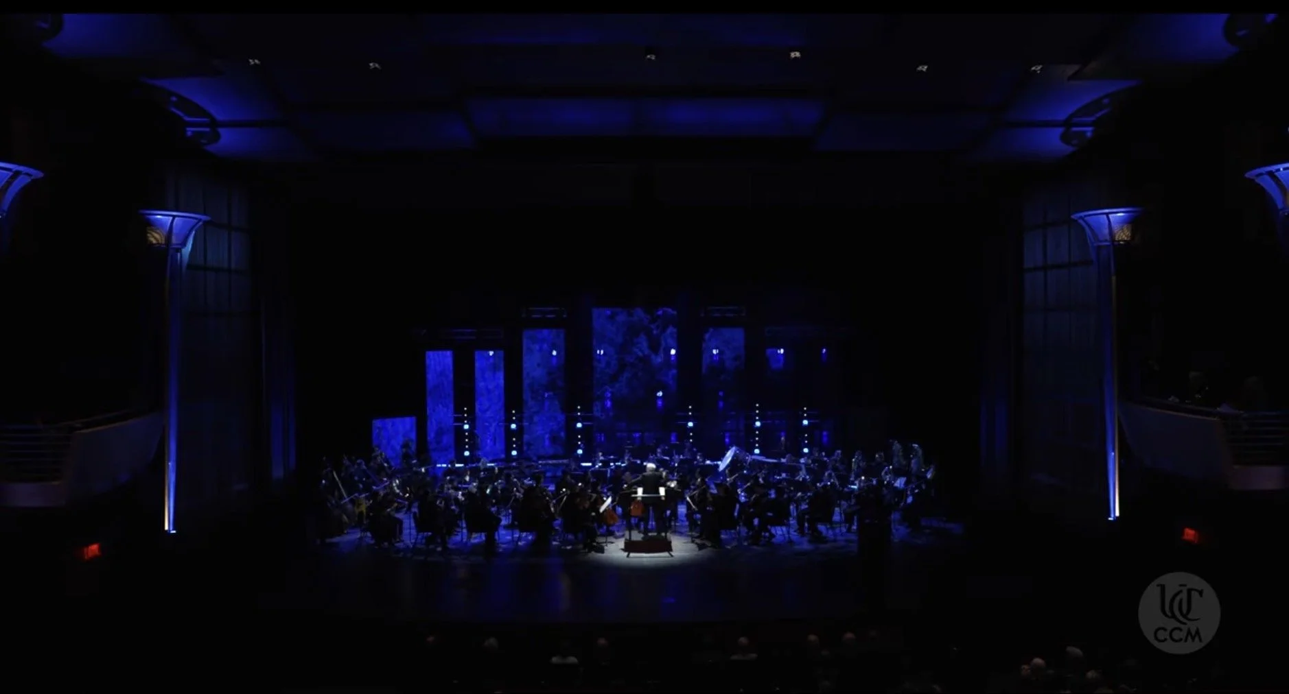 An orchestra performing on a dimly lit stage with blue lighting and abstract blue backdrop panels.