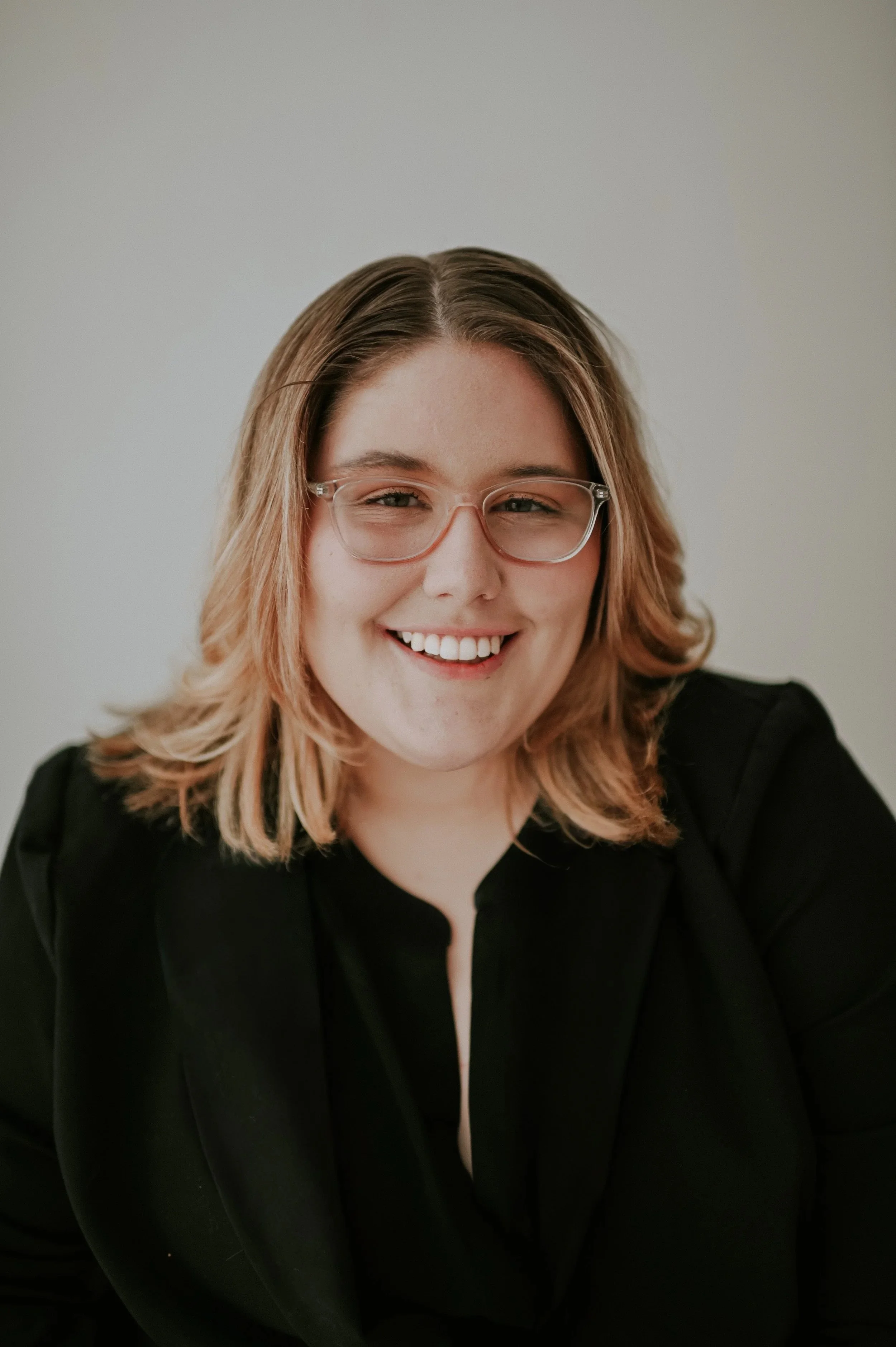 Close-up of a smiling woman with shoulder-length wavy blonde hair, wearing glasses and a black blazer against a plain background.