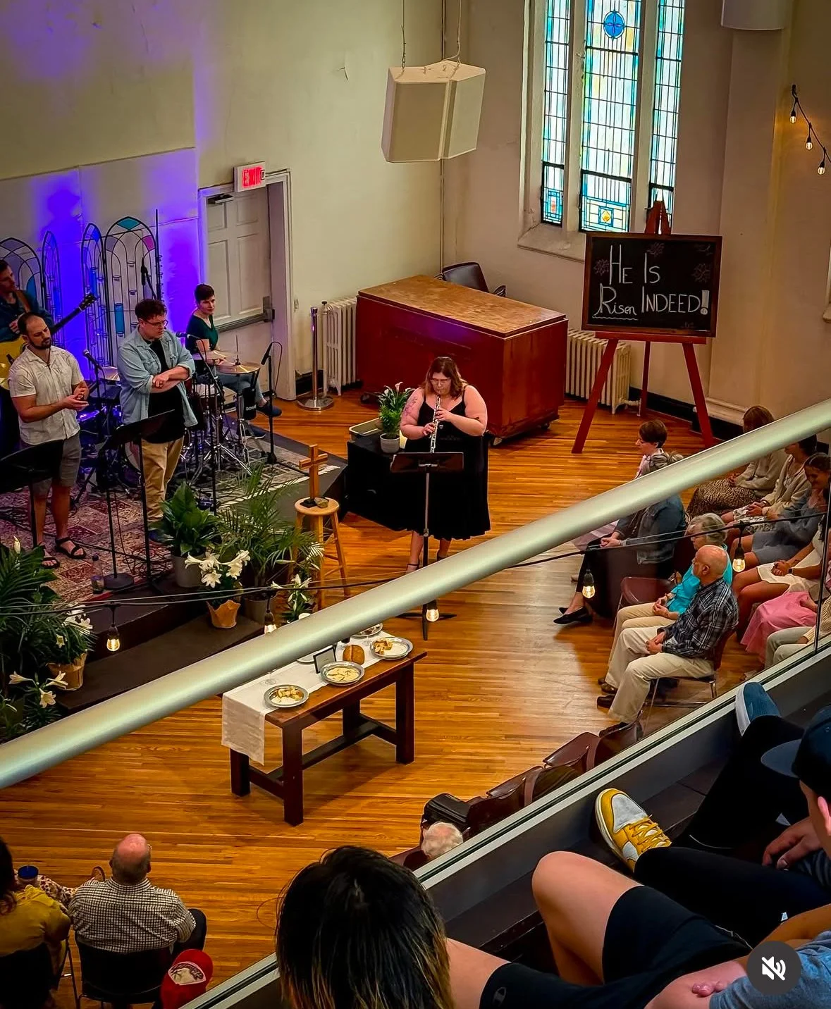 People performing and watching a live music performance inside a church or concert hall with stained glass windows, a blackboard with 'He Is Risen INDEED!' written on it, and audience seating.