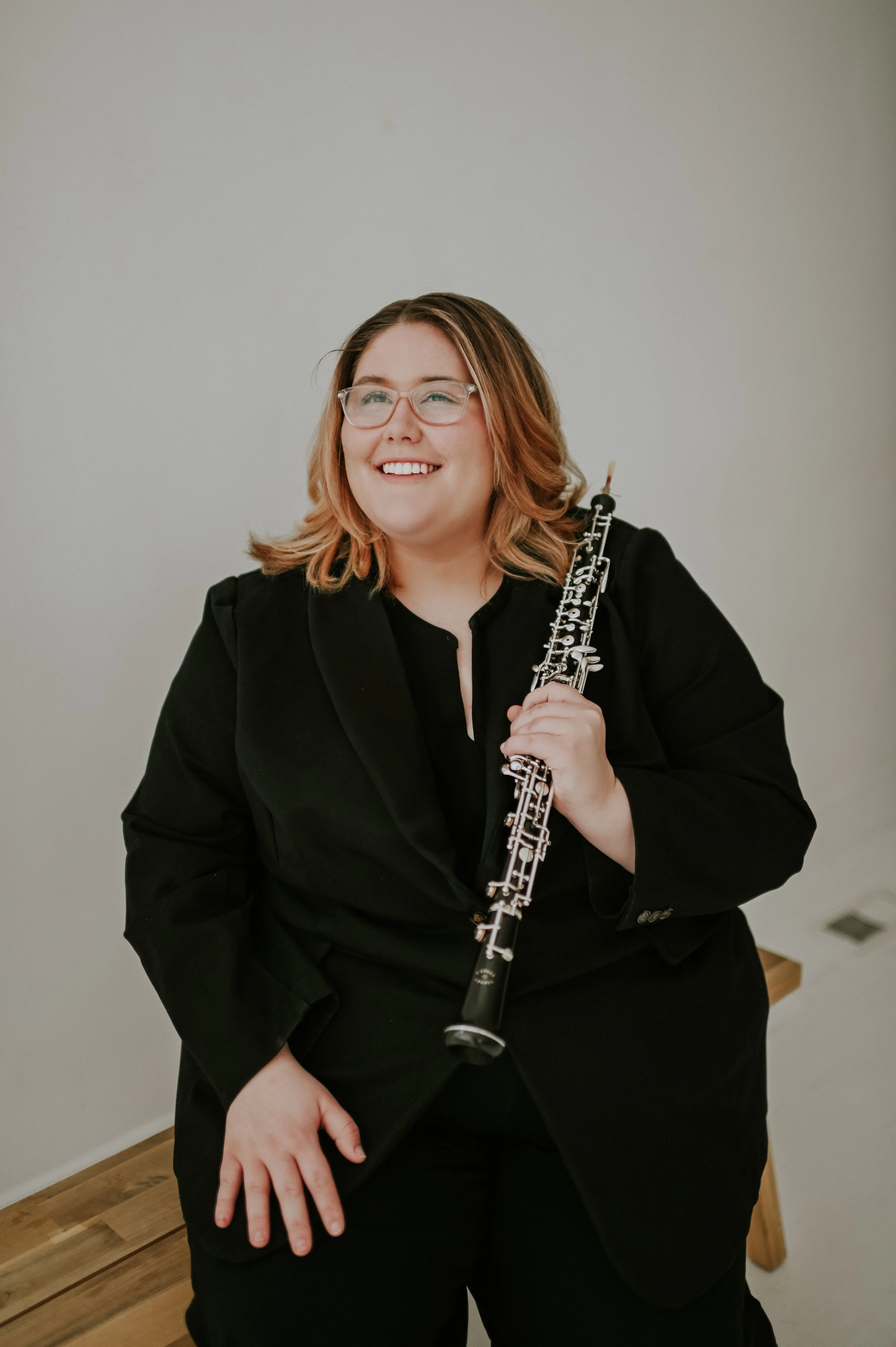 A woman with shoulder-length wavy hair, wearing glasses and a black blazer, sitting on a wooden bench, holding an oboe, smiling, in a room with a plain off-white wall.