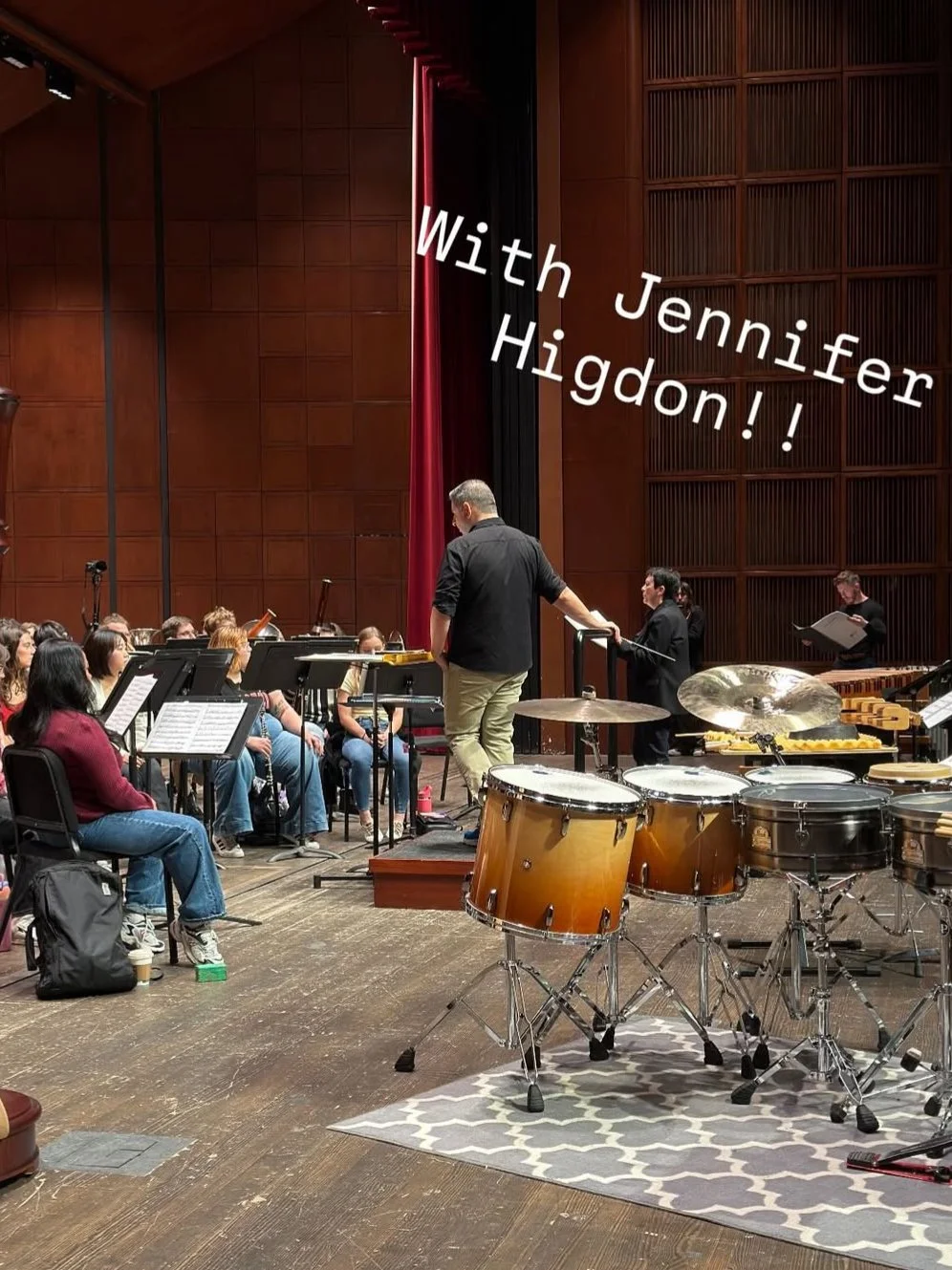 An orchestra rehearsal or concert in a wooden auditorium, with musicians seated and a conductor standing on a podium. Music stands are in front of the musicians, and percussion instruments, including drums, are visible in the foreground. A person is reading sheet music at a marimba or xylophone. Text overlay says 'With Jennifer Higdon!!'