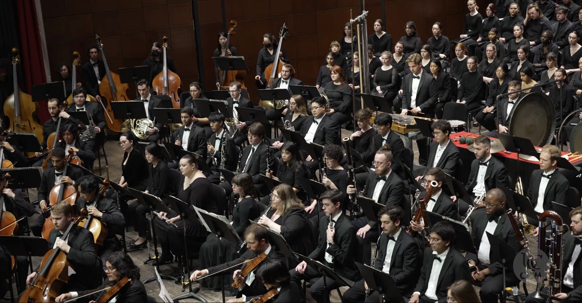 An orchestra performing on stage with musicians playing string, wind, and percussion instruments, dressed in formal black attire.